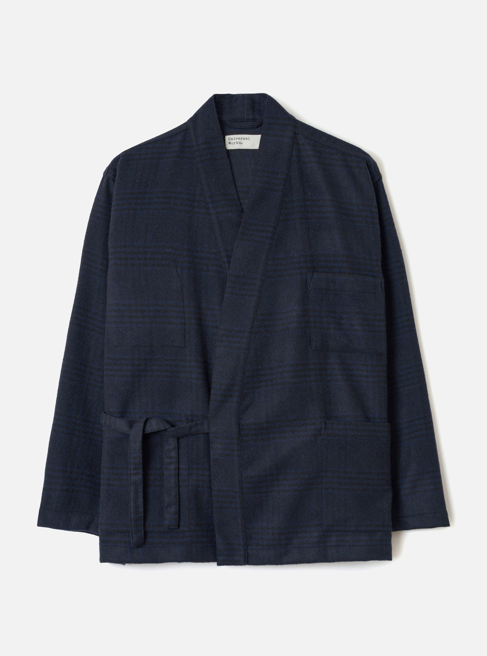Navy wool mix Kyoto work jacket in a tonal glen plaid pattern, flat lay on a light grey backdrop. The open-front jacket features a shawl collar, two large patch pockets at the hip, a chest patch pocket, and a self-tie belt at the waist. Long sleeves with plain cuffs. Universal Works woven label at the neckline.