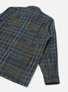 Flat lay of an olive shadows check fatigue shirt on a white studio surface, shot from above at an angle. The back panel and one long sleeve are visible, showing a brushed flannel fabric in an olive, navy, grey and blue check pattern. The buttoned cuff features two dark buttons.