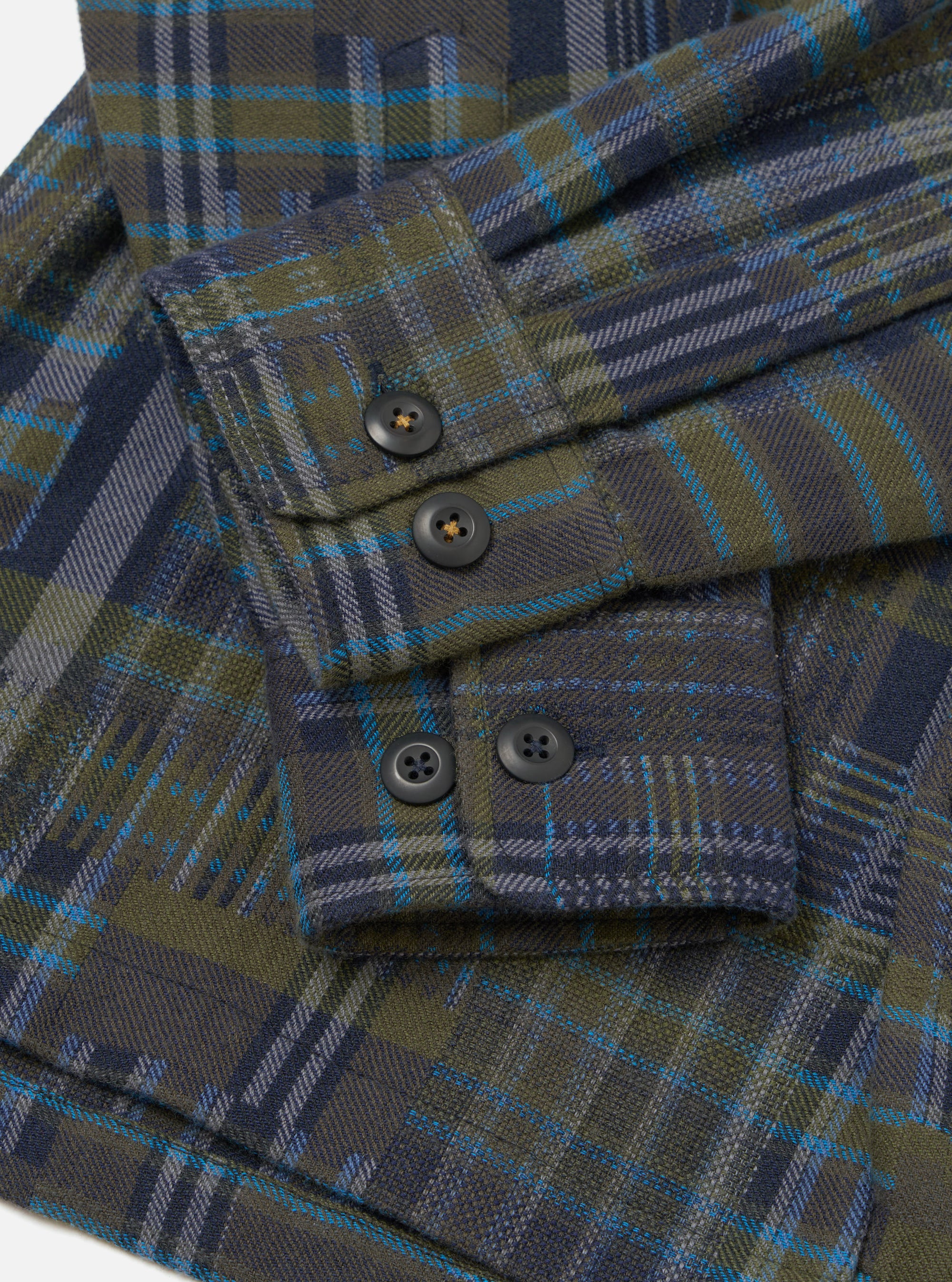 Close-up flat lay of an olive shadows check fatigue shirt in brushed flannel. The shot focuses on the sleeve and cuff, showing four dark horn-style buttons: two on the placket and two on the buttoned cuff. The plaid weave combines olive, navy, grey and teal on a white studio backdrop.