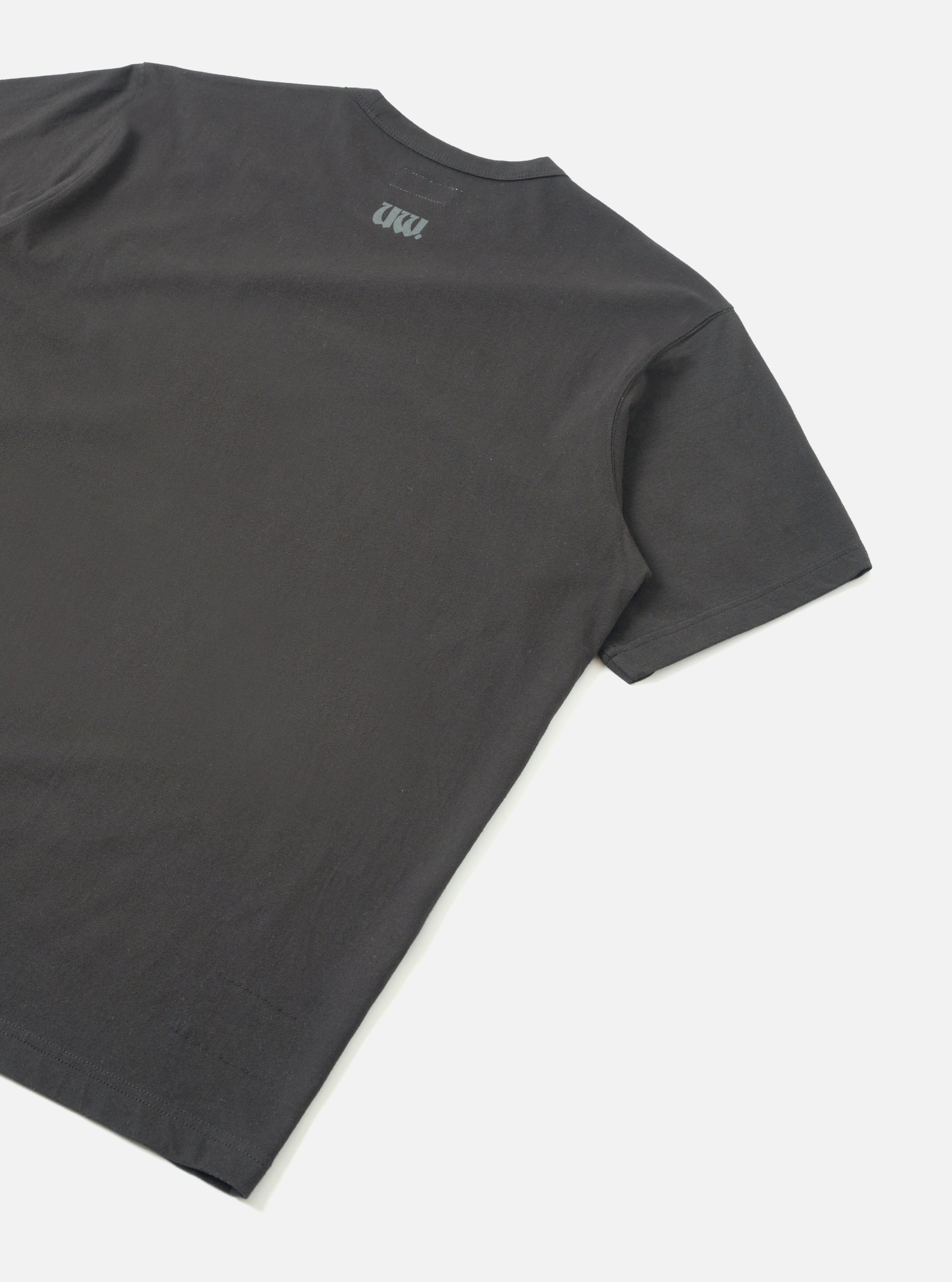Flat lay of a black single jersey t-shirt shot from behind on a light grey seamless backdrop. The back panel is plain with a small tonal UW logo print near the neckline. Smooth jersey fabric with a clean crew neck and short sleeves with hemmed cuffs visible.