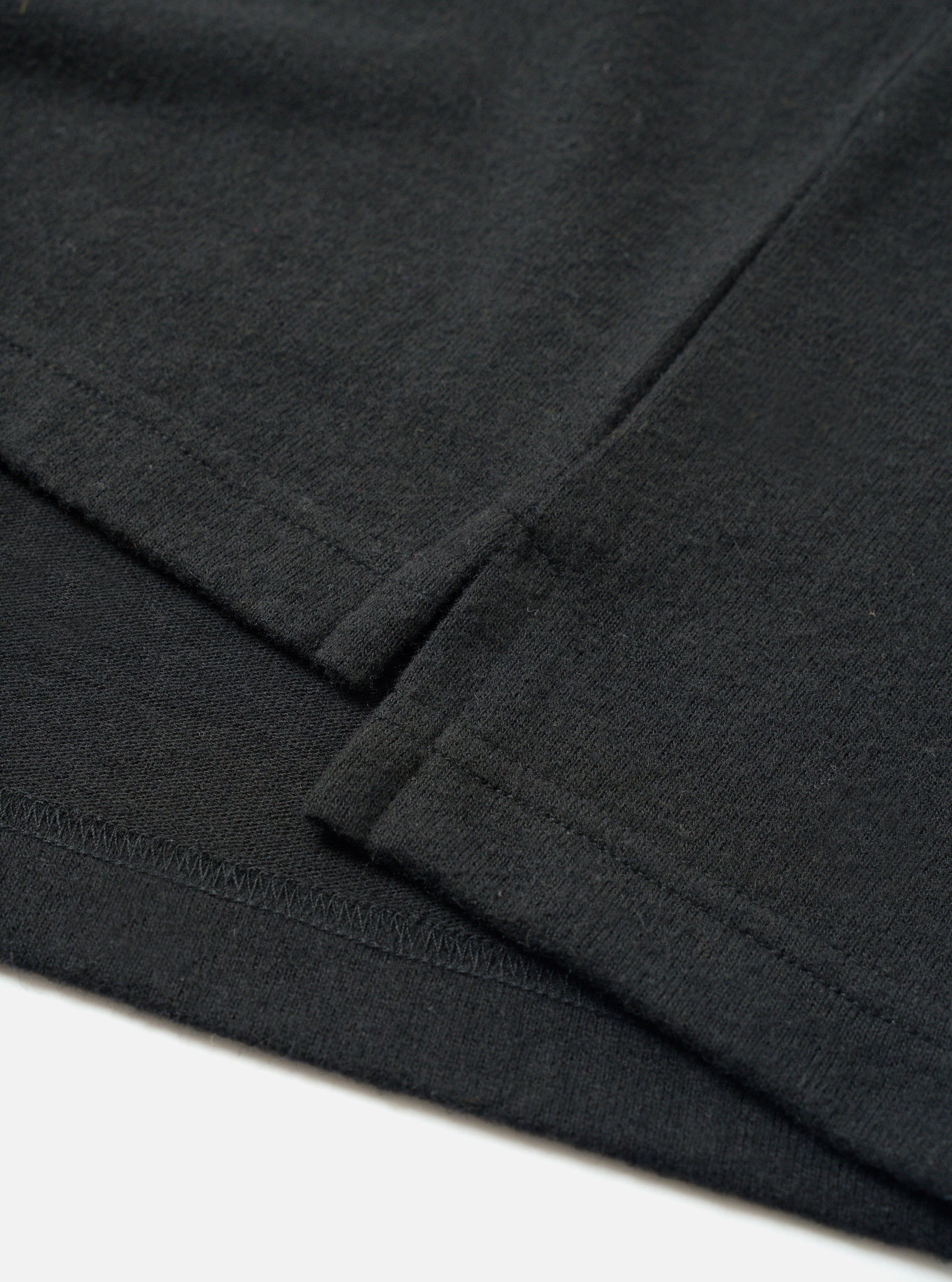 Close-up flat lay detail of the black recycled wool mix long-sleeve pocket tee. The shot focuses on the chest patch pocket and sleeve cuff, showing the folded ribbed cuff edge with visible topstitching and a zigzag overlock seam. The fine-knit jersey fabric has a subtle heathered texture. White surface background.