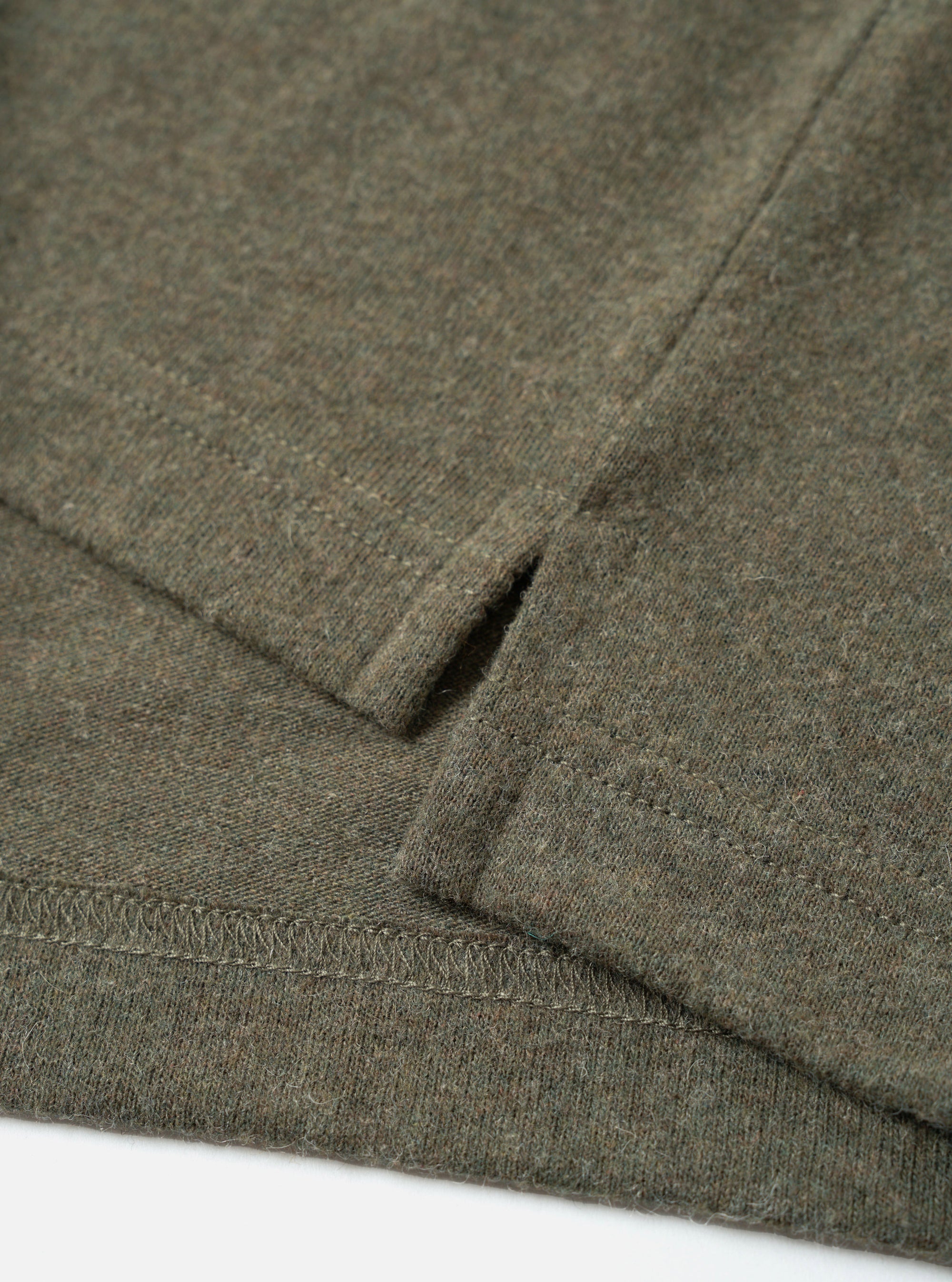 Close-up flat lay of an olive marl recycled wool mix long-sleeve pocket tee on a white surface. The shot focuses on the lower hem and cuff, showing a ribbed hem with overlock stitching, a folded ribbed cuff, and a small welt thumb loop detail. The heathered fabric has a soft, lightly textured finish.