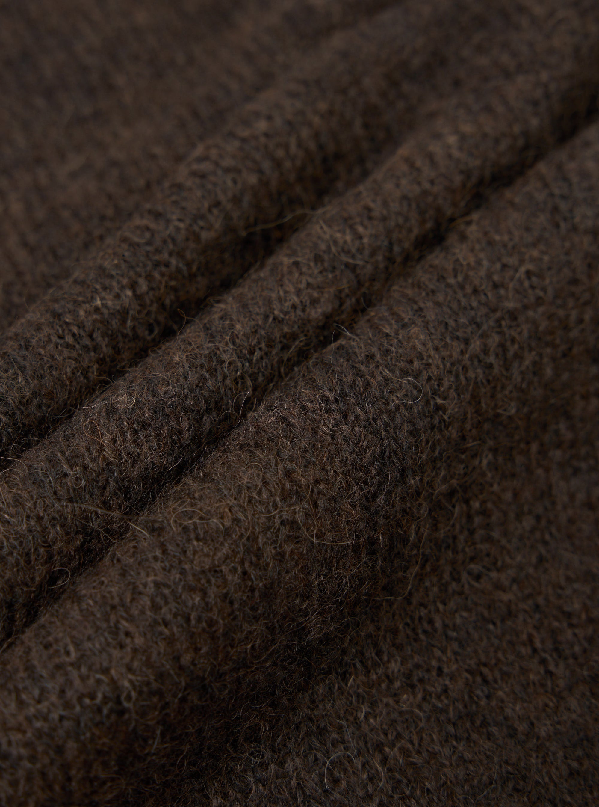 Close-up detail shot of the brown alpaca knit fabric of the David Cardigan, showing soft, fluffy fibres with a hairy, textured surface. Three gentle folds run diagonally across the frame, highlighting the fabric's depth and loft. The dark brown marl tone shows subtle variation throughout.