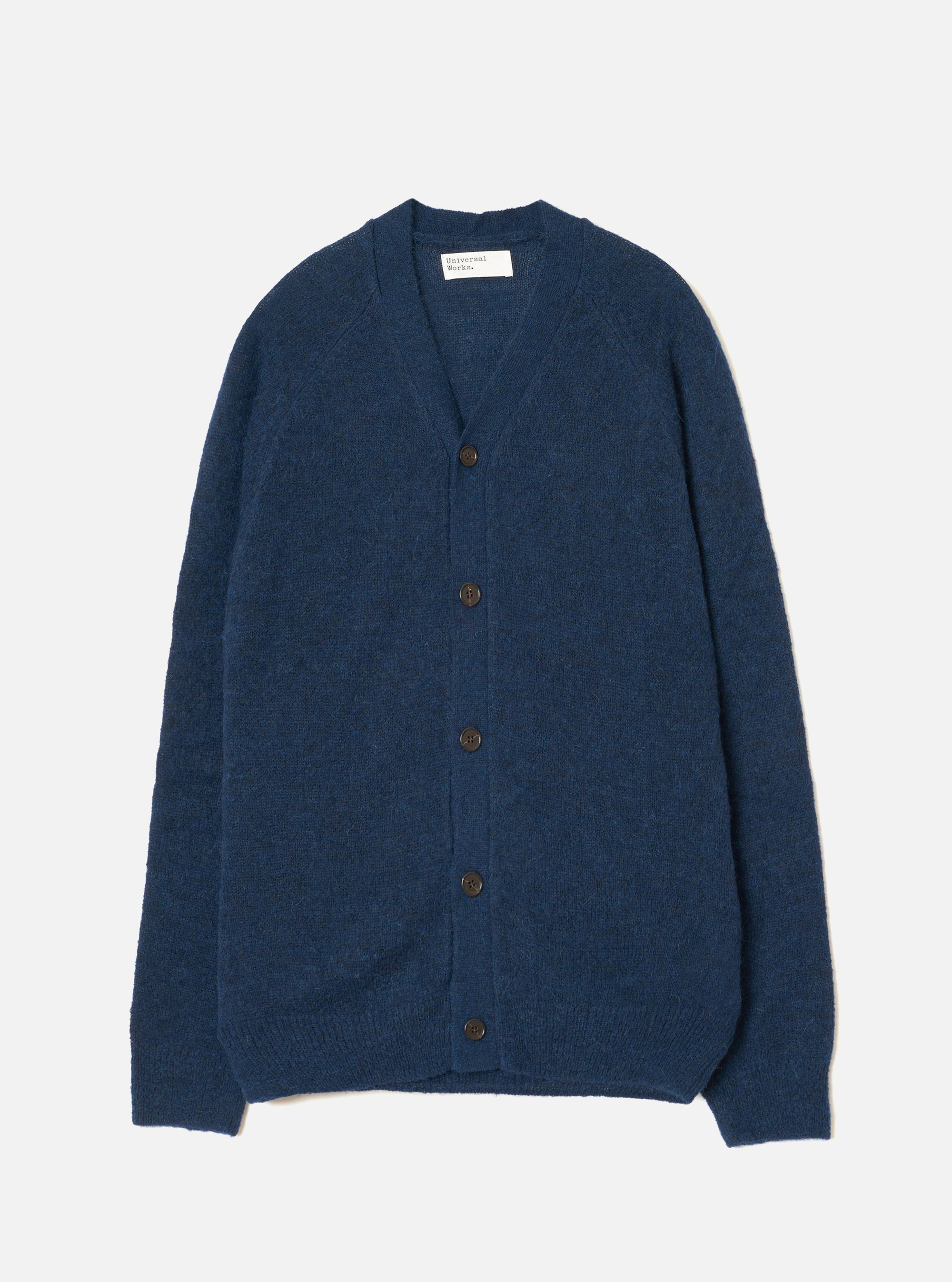 Navy alpaca knit cardigan laid flat on a light grey seamless backdrop. Features a V-neckline, raglan sleeves, and five dark brown buttons down the centre front. Ribbed cuffs and hem visible. The fluffy, brushed texture of the alpaca knit is evident across the body. A Universal Works woven label sits at the inner neck.