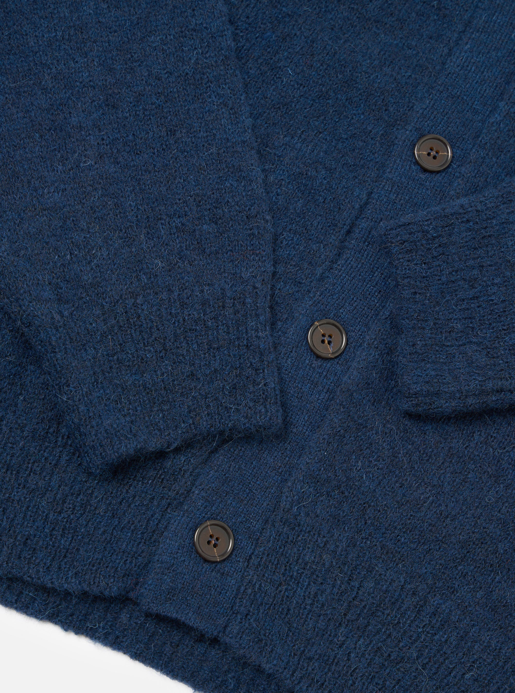 Close-up flat lay of the navy alpaca knit David cardigan on a white surface, showing the button placket with three dark four-hole buttons, a patch chest pocket, and the ribbed cuff of one sleeve. The fabric has a soft, fluffy texture with a fine knit construction.