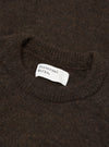 Close-up flat lay of a brown alpaca knit crew neck sweater. The ribbed crew neckline is visible at the top of the frame, with a white woven Universal Works brand label stitched inside. The fluffy, hairy texture of the alpaca knit fabric fills the frame. Shot on a plain surface.