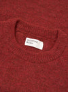 Close-up flat lay of the Universal Works deep orange alpaca knit crew neck, showing the ribbed crewneck collar and soft, fluffy knit texture in a dark burnt-orange marl. A white woven Universal Works label is visible inside the collar. Shot on a plain white surface.