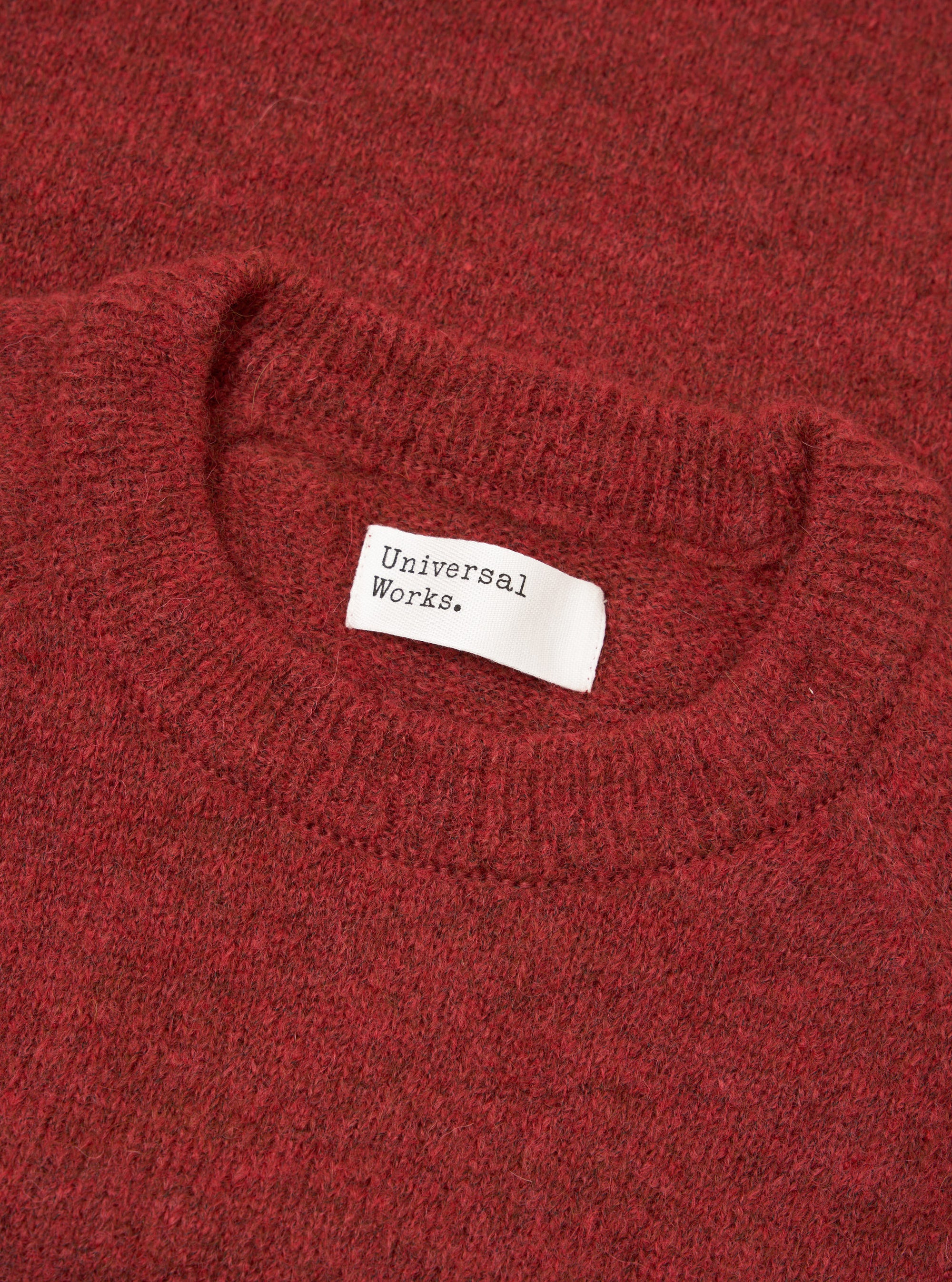 Close-up flat lay of the Universal Works deep orange alpaca knit crew neck, showing the ribbed crewneck collar and soft, fluffy knit texture in a dark burnt-orange marl. A white woven Universal Works label is visible inside the collar. Shot on a plain white surface.