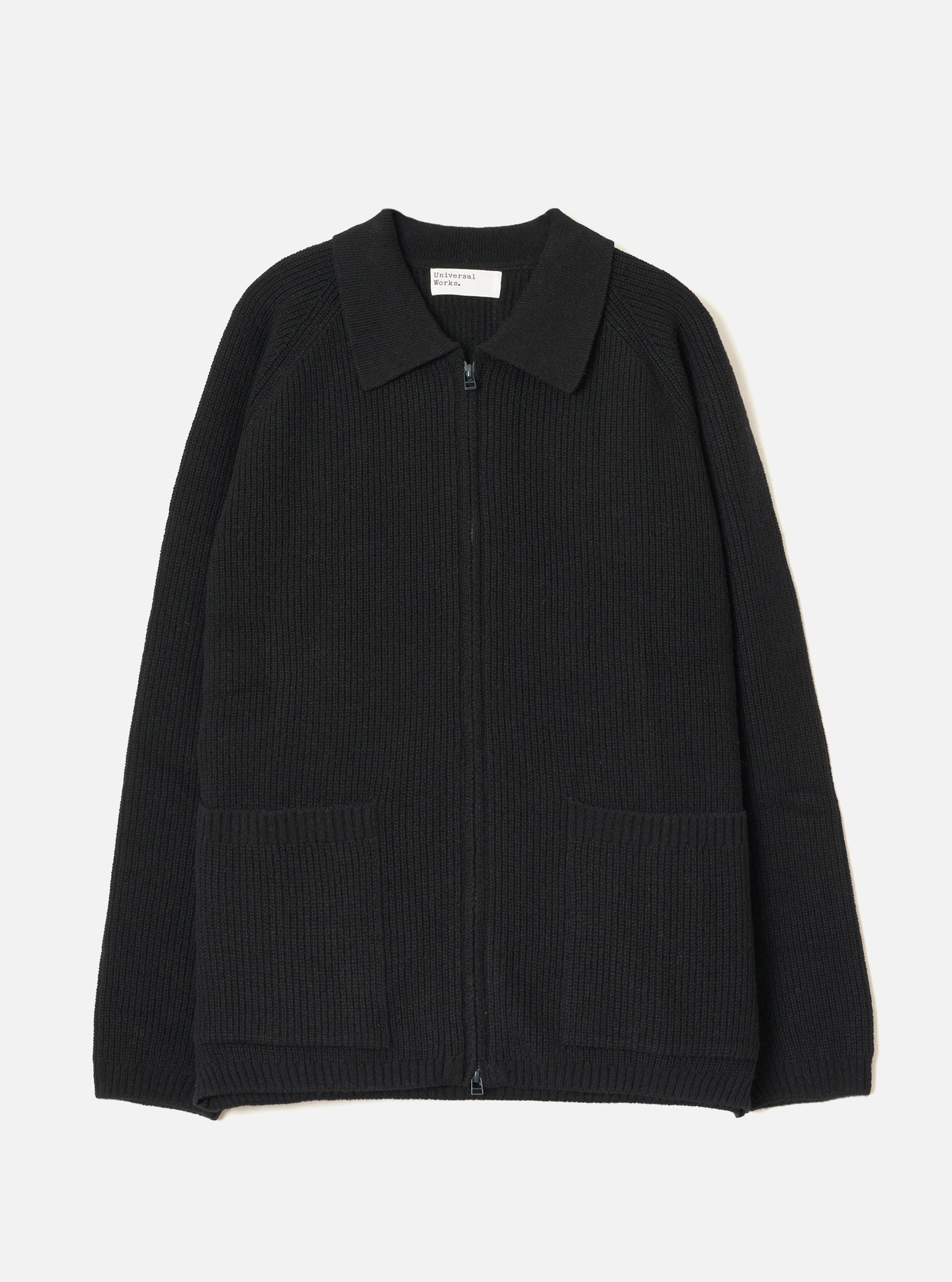 Black ribbed-knit zip cardigan laid flat on a light grey seamless backdrop. Features a spread collar, full-length zip fastening, raglan sleeves, two large patch pockets at the hem, and a wide ribbed waistband. Universal Works woven label visible at the inner collar. No model or mannequin present.