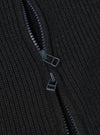 Close-up detail of the black eco wool zip cardigan showing a two-way zip partially open. Two dark gunmetal zip pullers are visible against the chunky ribbed knit fabric. The zip teeth and tape are tonal dark grey, blending with the wool texture. No background is visible.