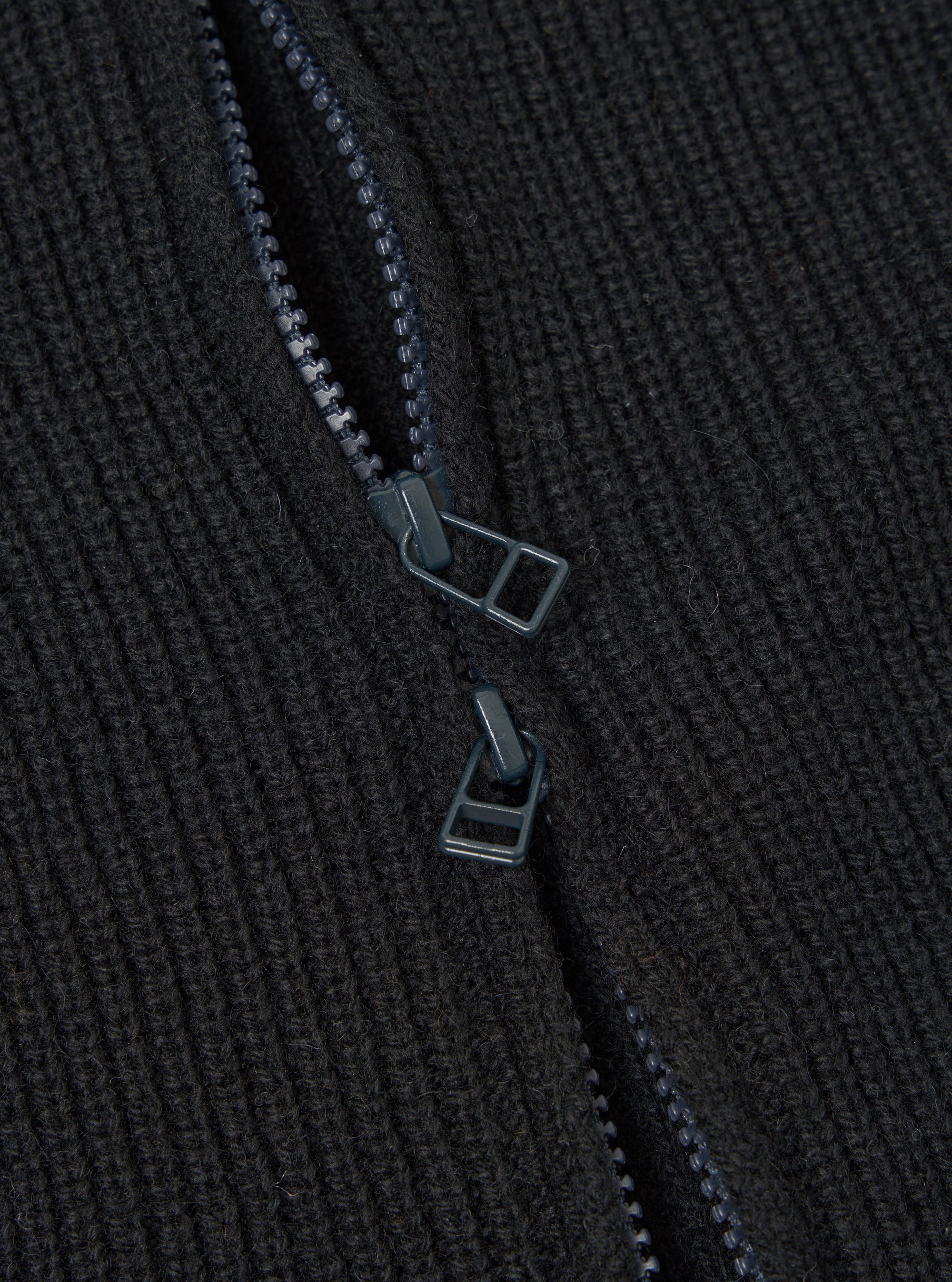 Close-up detail of the black eco wool zip cardigan showing a two-way zip partially open. Two dark gunmetal zip pullers are visible against the chunky ribbed knit fabric. The zip teeth and tape are tonal dark grey, blending with the wool texture. No background is visible.