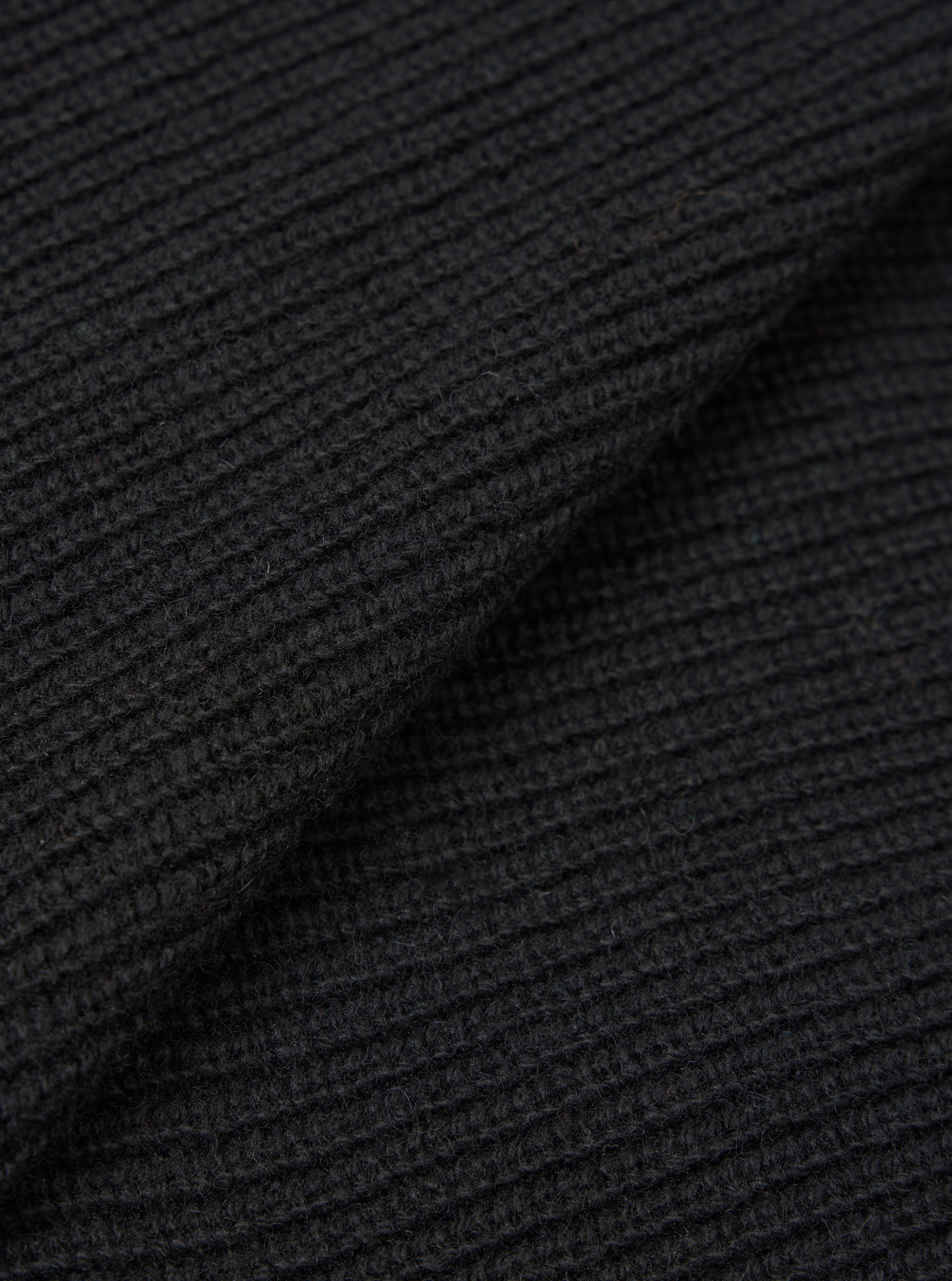 Close-up of black eco wool fabric showing a chunky waffle-knit texture with horizontal ribbing. The material is folded to reveal depth and dimension in the knit structure. Individual yarn fibres are visible across the surface.
