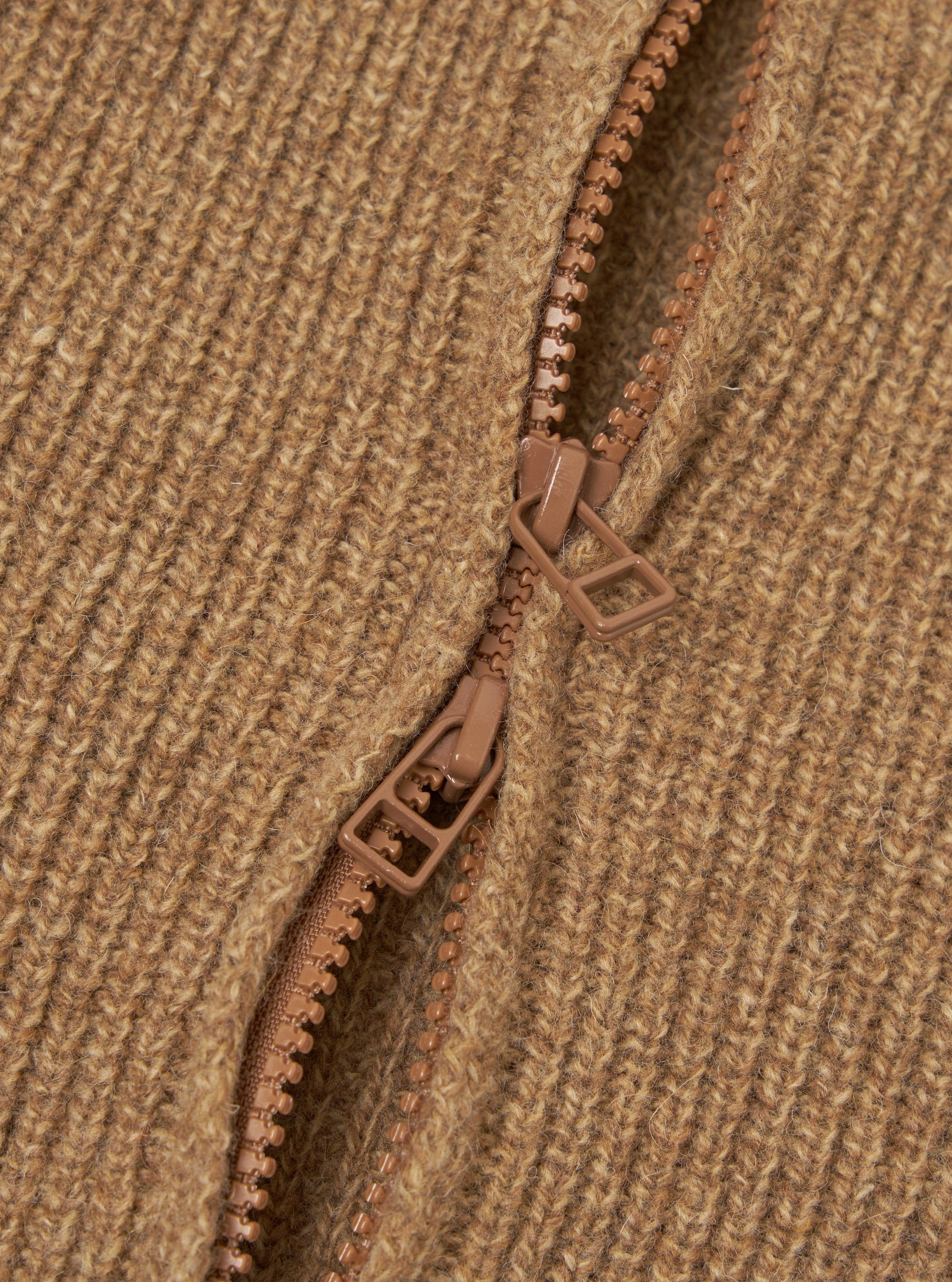 Close-up detail of the tawny eco wool zip cardigan showing a two-way brown zip partially open against chunky ribbed knit fabric. The zip pulls and teeth are tonal brown plastic. The hairy, textured wool-blend rib is visible across the full frame.