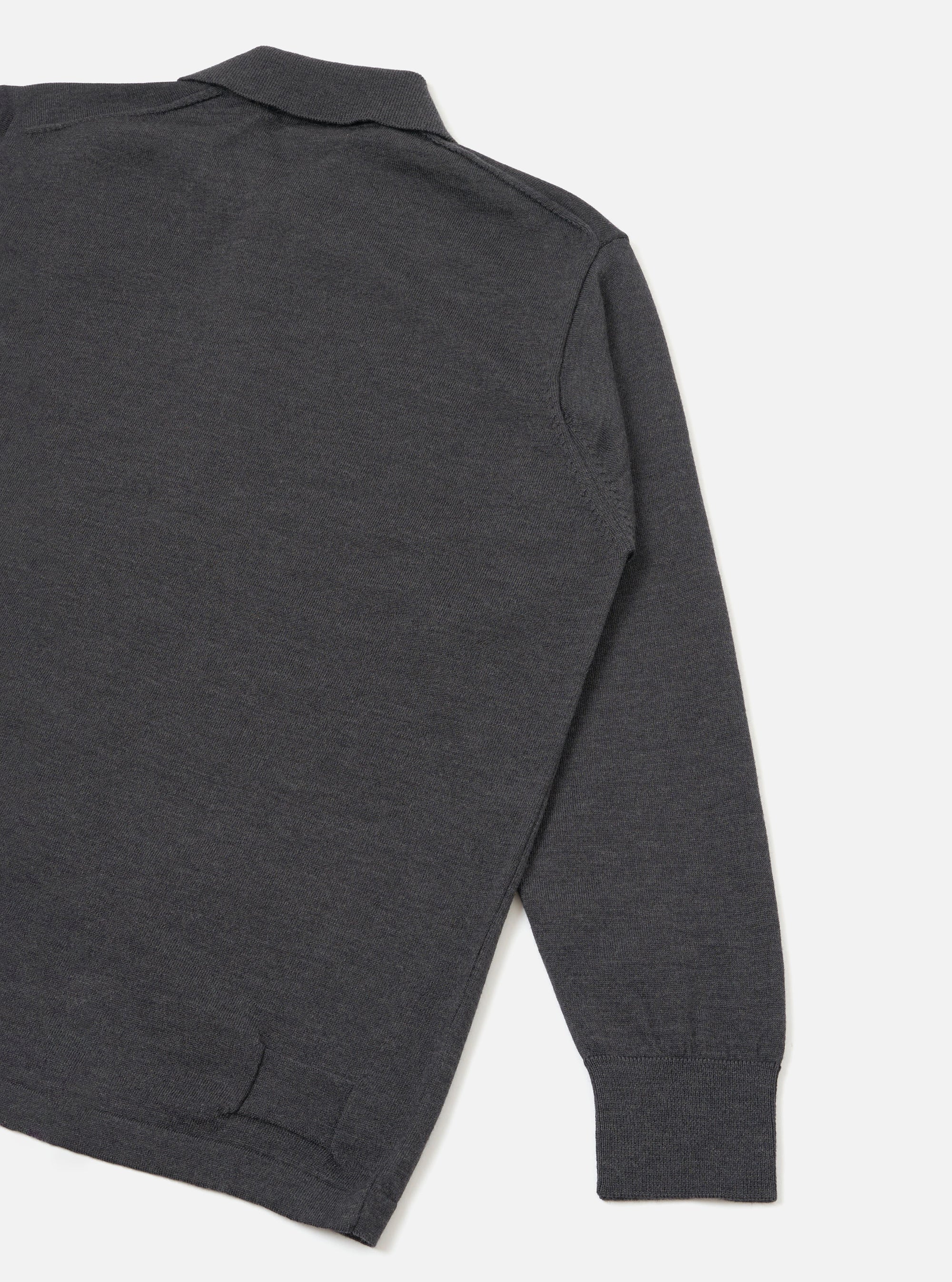 Close-up flat lay of a charcoal merino long-sleeve pullover knit shirt on a light grey seamless backdrop. The shot shows the upper back, shoulder and full length of one sleeve. A spread collar is visible at the top, the fabric has a fine knit texture, and a ribbed cuff finishes the sleeve. A partial hem vent is visible at the lower left.