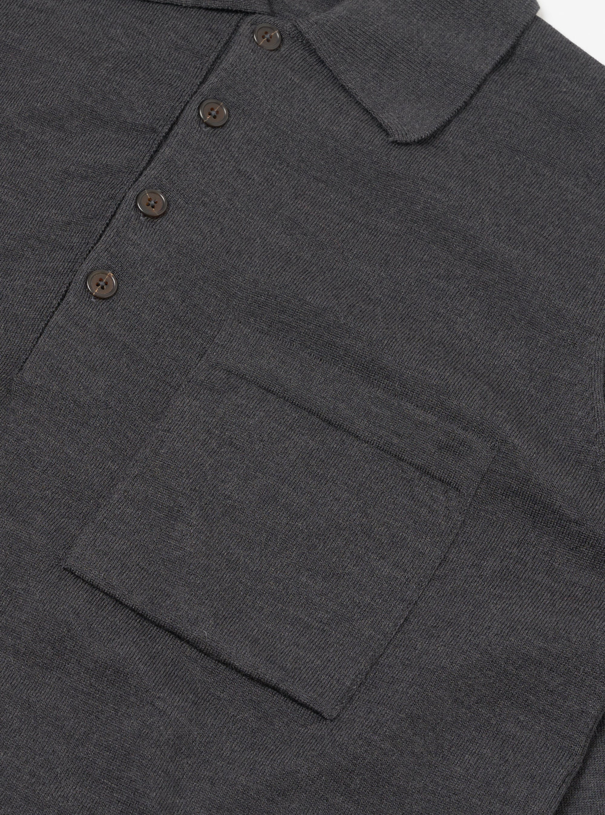 Close-up flat lay of a charcoal merino long-sleeve knit polo shirt on a white surface. The fine-knit fabric has a smooth, dense texture. Visible details include a spread collar, a four-button placket with dark tonal buttons, and a large patch chest pocket with a horizontal seam detail at the base.