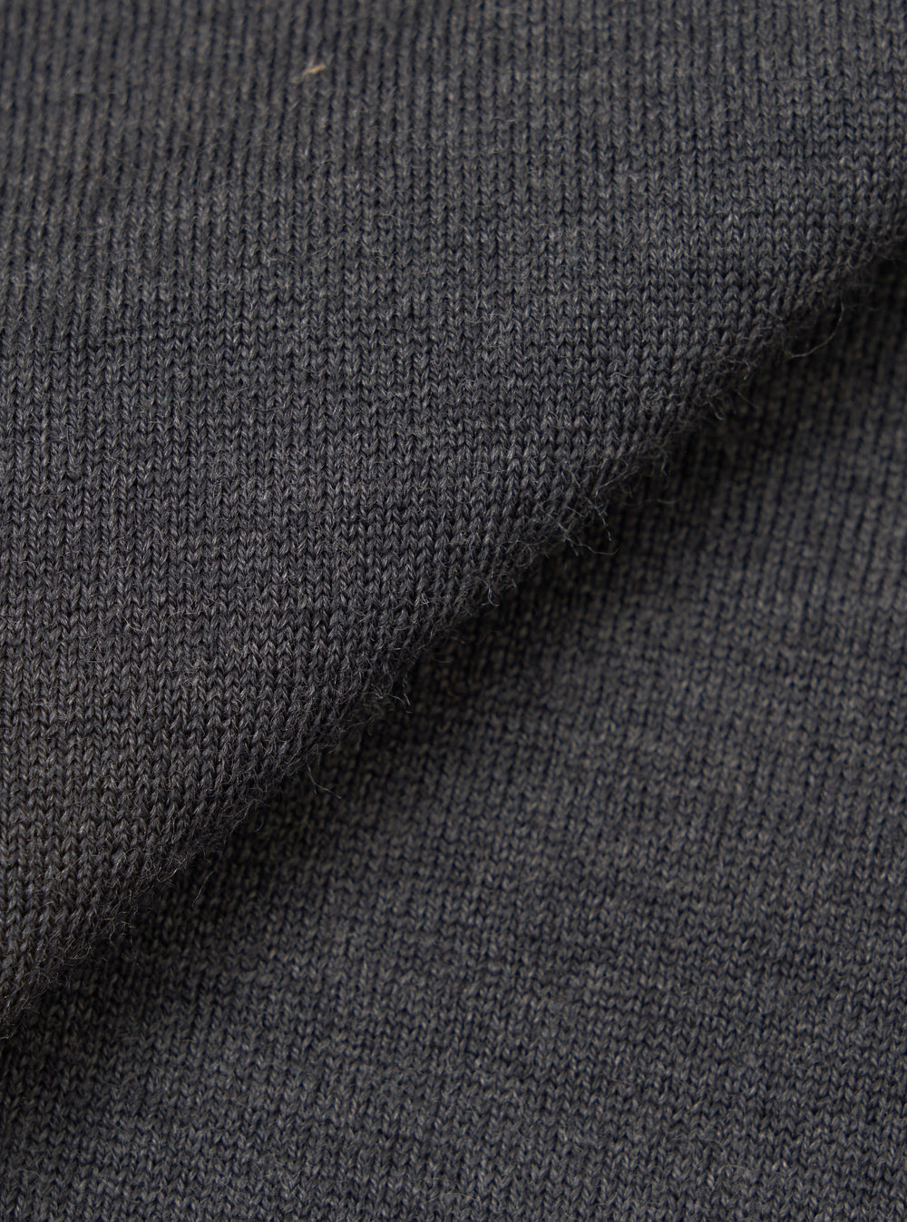 Close-up of the charcoal merino knit fabric, showing a fine, uniform jersey stitch with a soft, slightly textured surface. The dark grey tone has a subtle heathered quality. The fabric is folded to display drape and thickness against a plain white background.