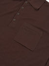 Close-up flat lay of an espresso brown merino knit polo shirt on a plain white surface. The ribbed knit collar, three-button placket with dark four-hole buttons, and a chest patch pocket are clearly visible. The fine knit texture is evident across the body.