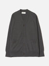 Charcoal merino cardigan laid flat on a light grey seamless backdrop. V-neckline with a woven Universal Works label inside. Four dark horn-effect buttons run down the front placket. Two patch pockets sit at the lower front. Ribbed cuffs and hem. Fine, smooth knit texture throughout. Full length, unbutoned view from the front.