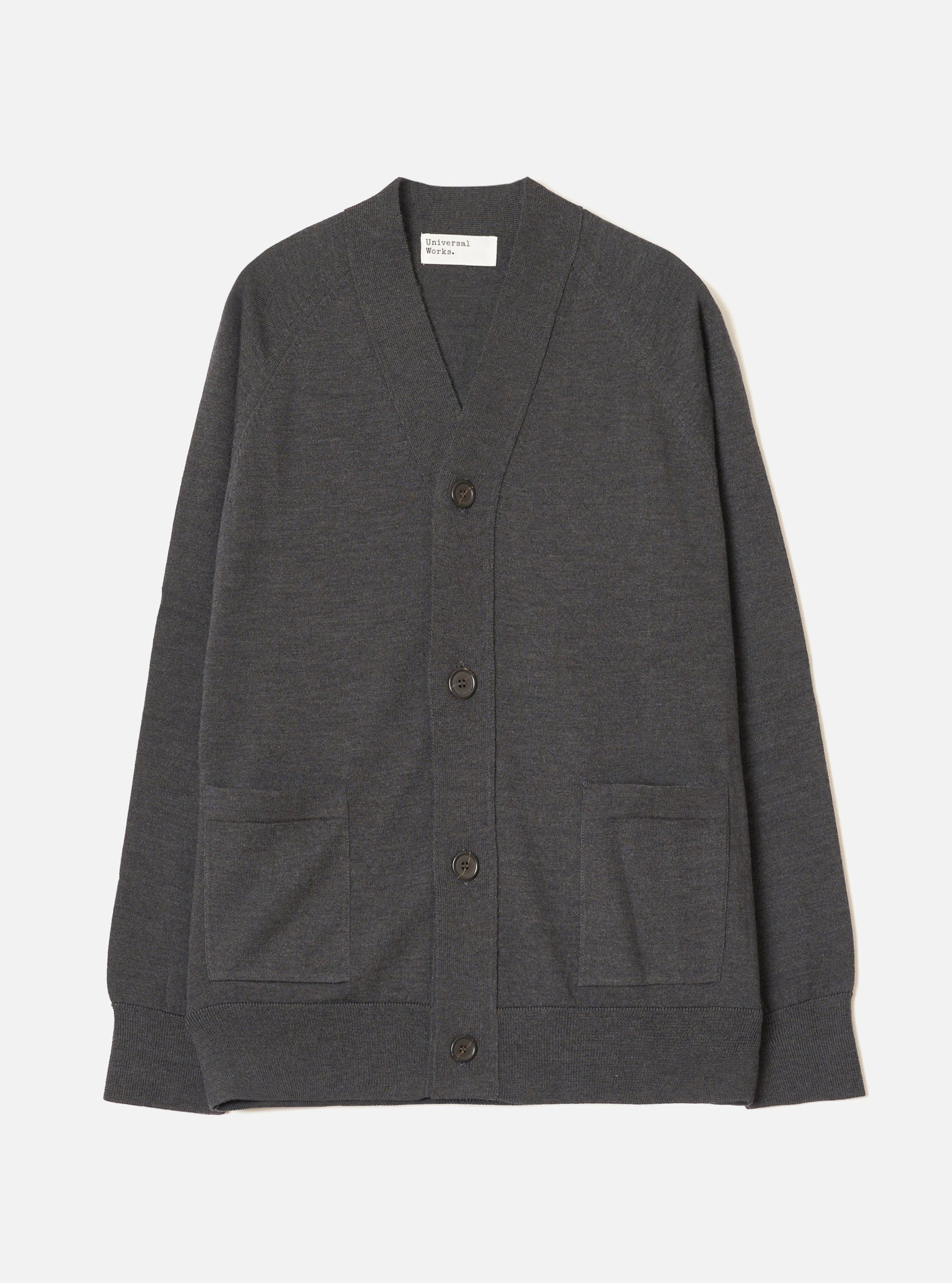 Charcoal merino cardigan laid flat on a light grey seamless backdrop. V-neckline with a woven Universal Works label inside. Four dark horn-effect buttons run down the front placket. Two patch pockets sit at the lower front. Ribbed cuffs and hem. Fine, smooth knit texture throughout. Full length, unbutoned view from the front.