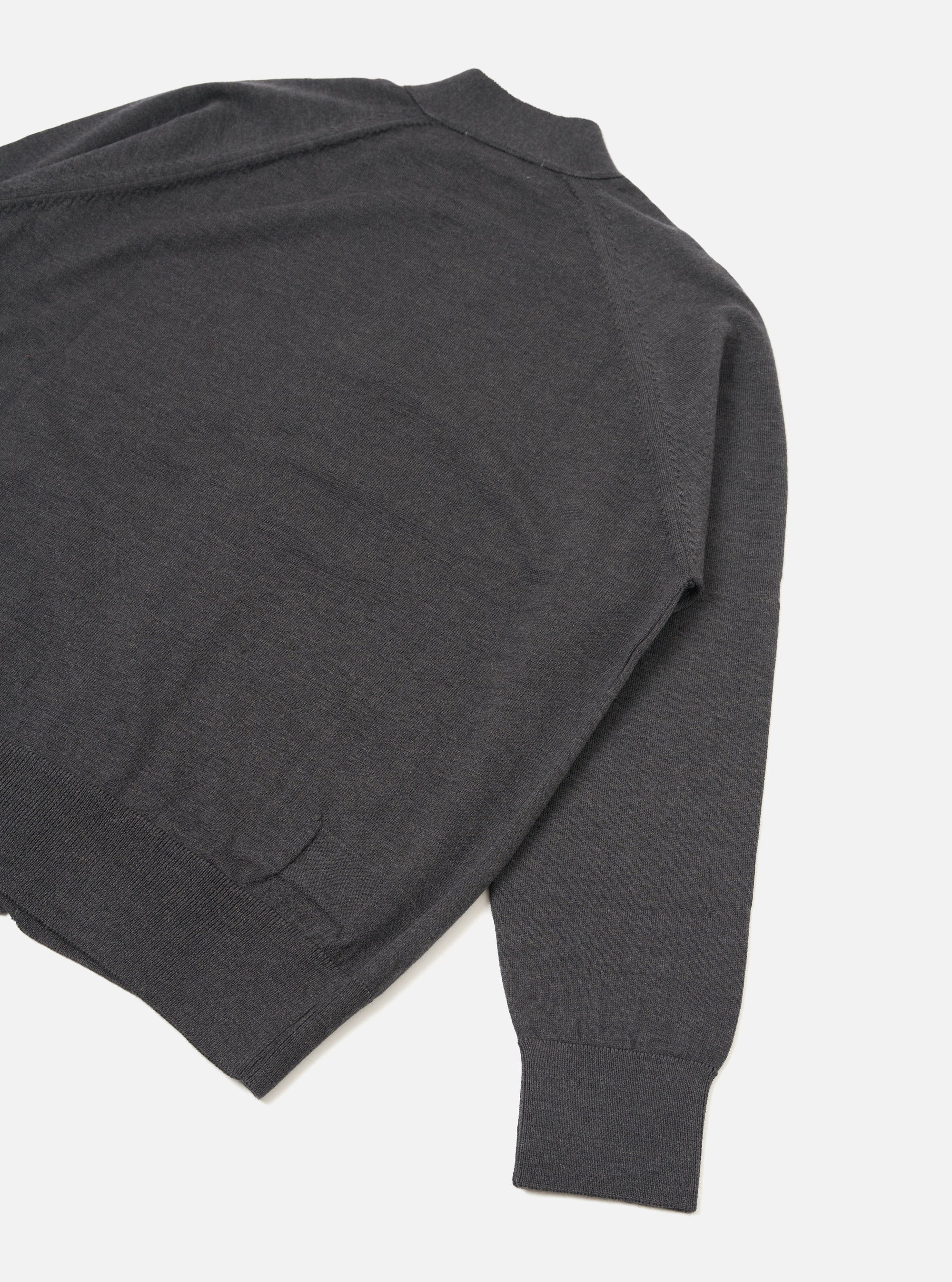 Flat lay of a charcoal merino loose cardigan on a white studio backdrop, shot from above at an angle. The fine-knit body shows a mock neck or collar at the top, a ribbed hem, and one full sleeve with a ribbed cuff. A seam detail runs along the sleeve join.
