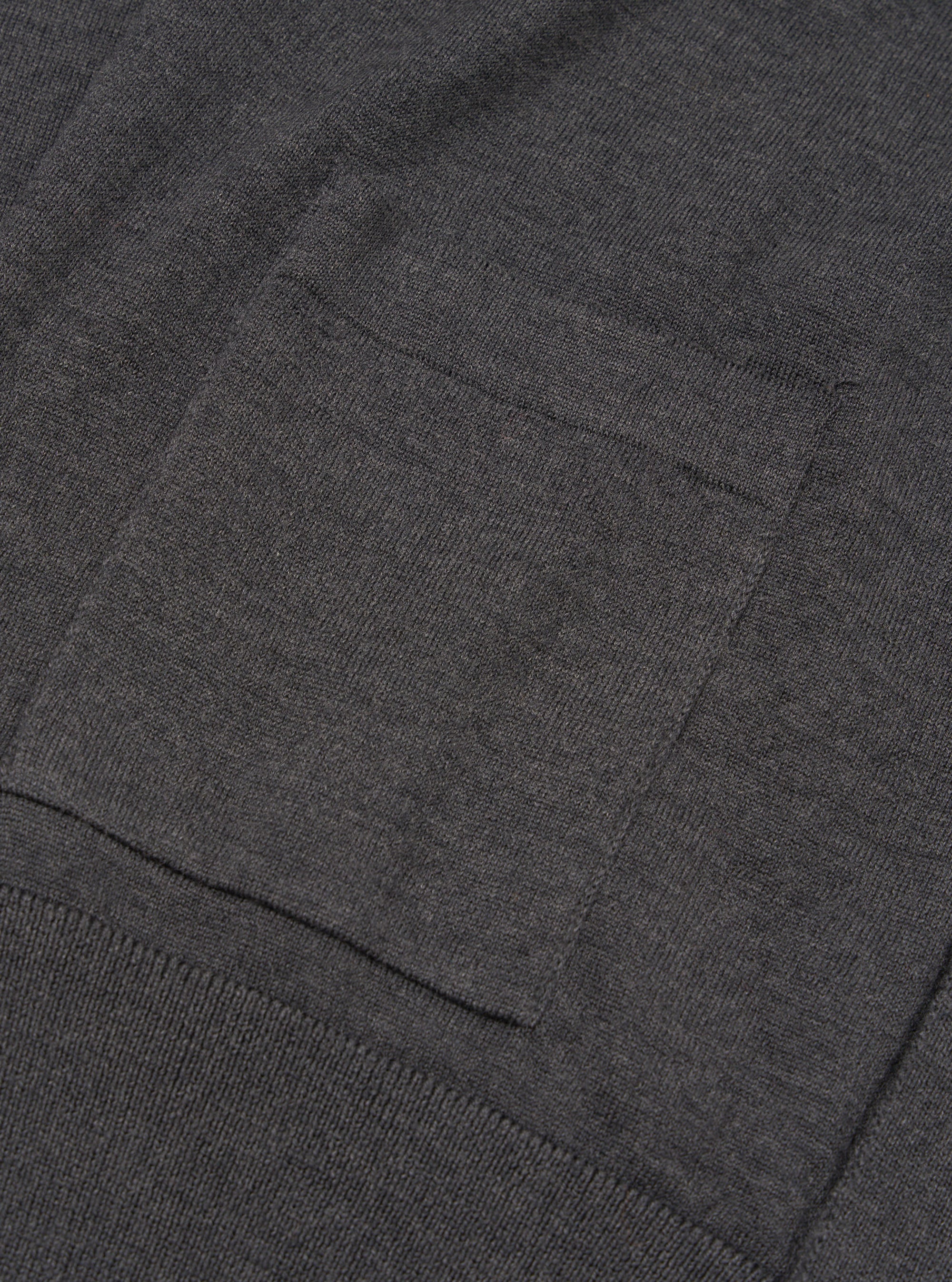 Close-up detail of a charcoal merino cardigan showing a patch chest pocket with neat stitched edges and a ribbed hem band below. The fine-knit merino fabric has a smooth, dense texture in a dark grey marl. Shot flat against a plain background.