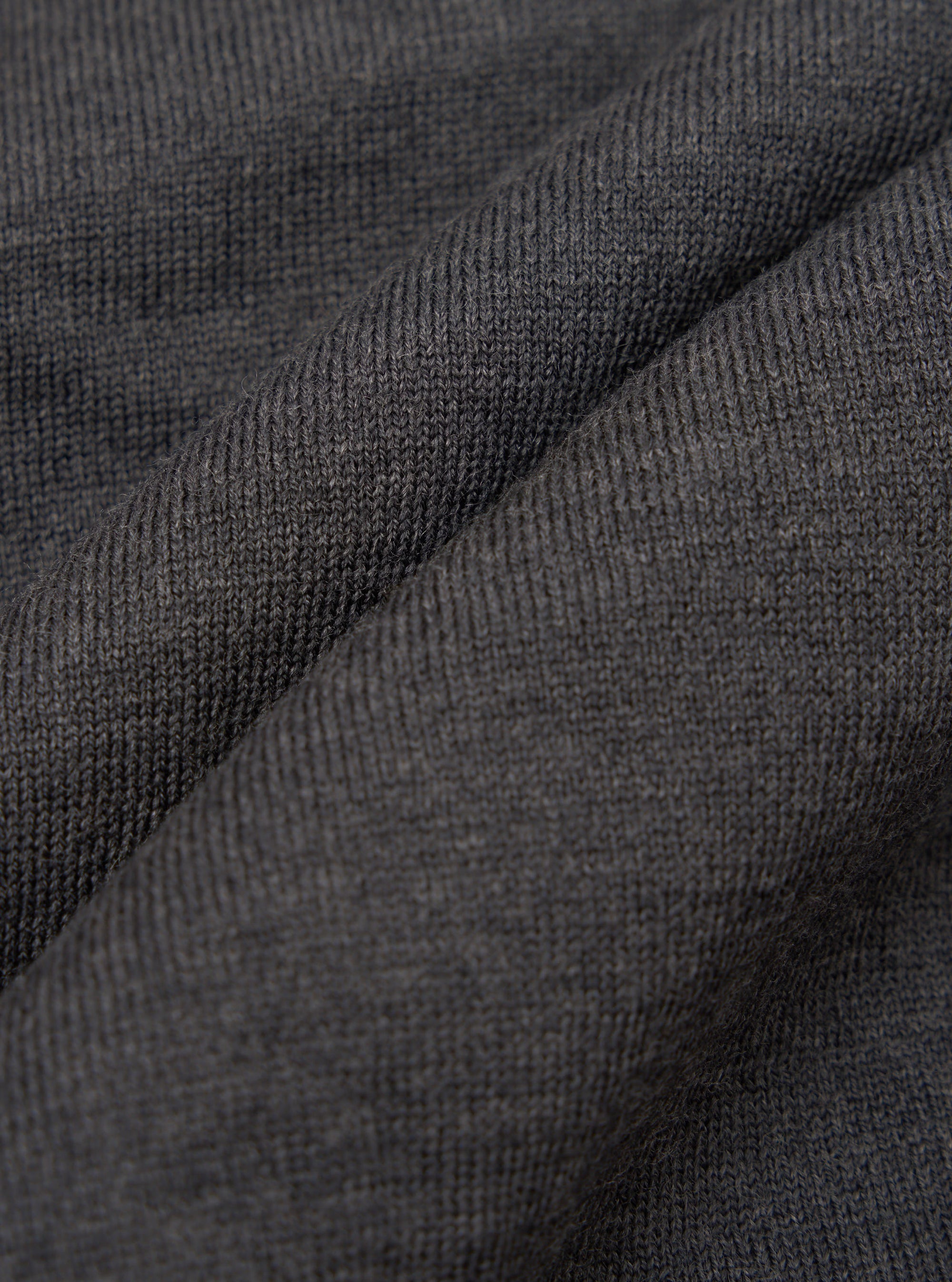 Close-up of charcoal merino knit fabric, showing a fine, even stocking stitch with a soft, slightly heathered texture. The fabric is gathered into soft folds, highlighting the drape and weight of the knit. No background is visible.