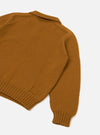 Gold Shetland wool sweater, flat lay on a white studio surface, shot from above at an angle showing the back and one sleeve. Chunky knit texture throughout, ribbed hem and cuff visible, spread collar at the top. A small woven patch detail sits near the hem.