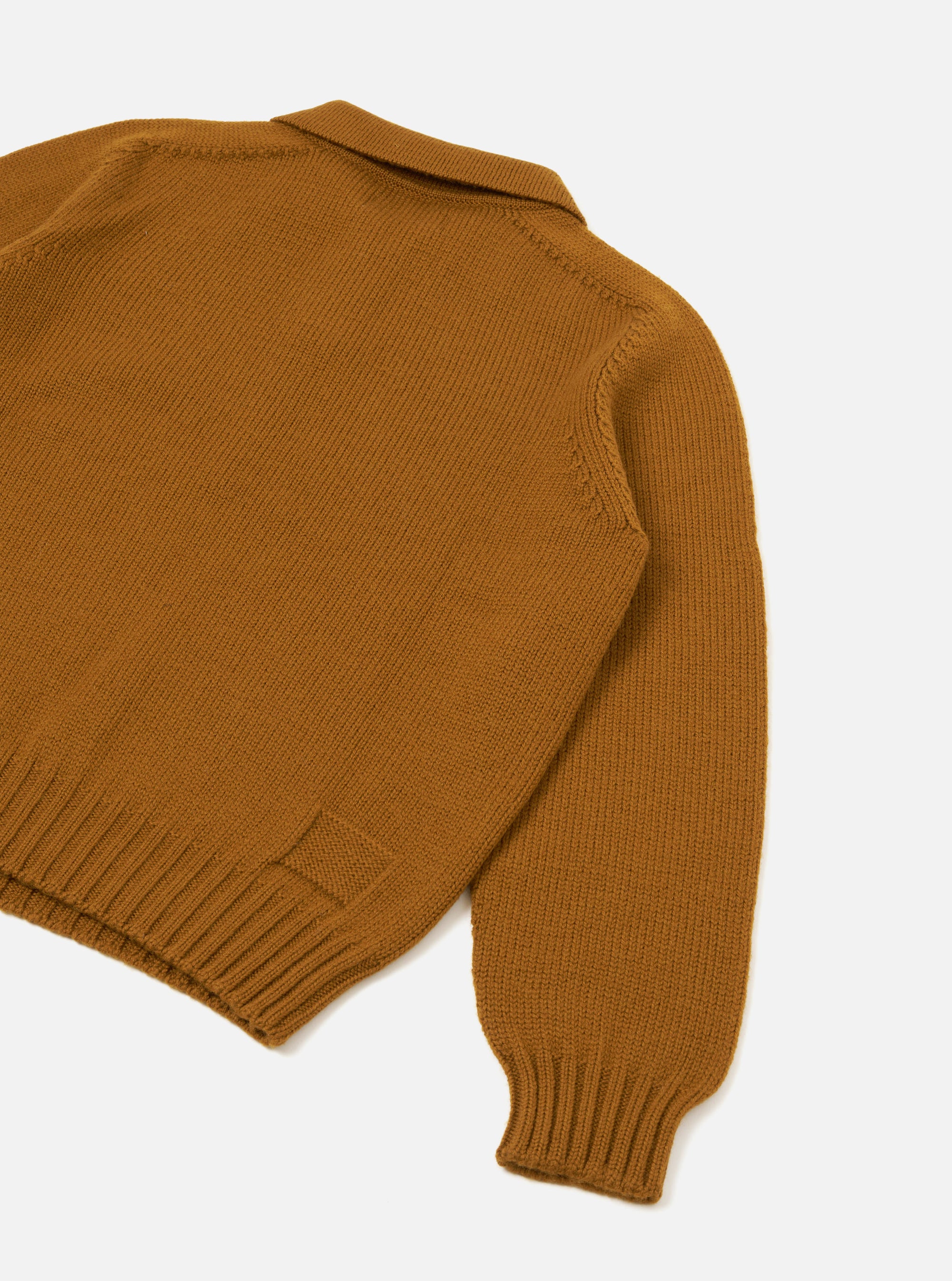 Gold Shetland wool sweater, flat lay on a white studio surface, shot from above at an angle showing the back and one sleeve. Chunky knit texture throughout, ribbed hem and cuff visible, spread collar at the top. A small woven patch detail sits near the hem.