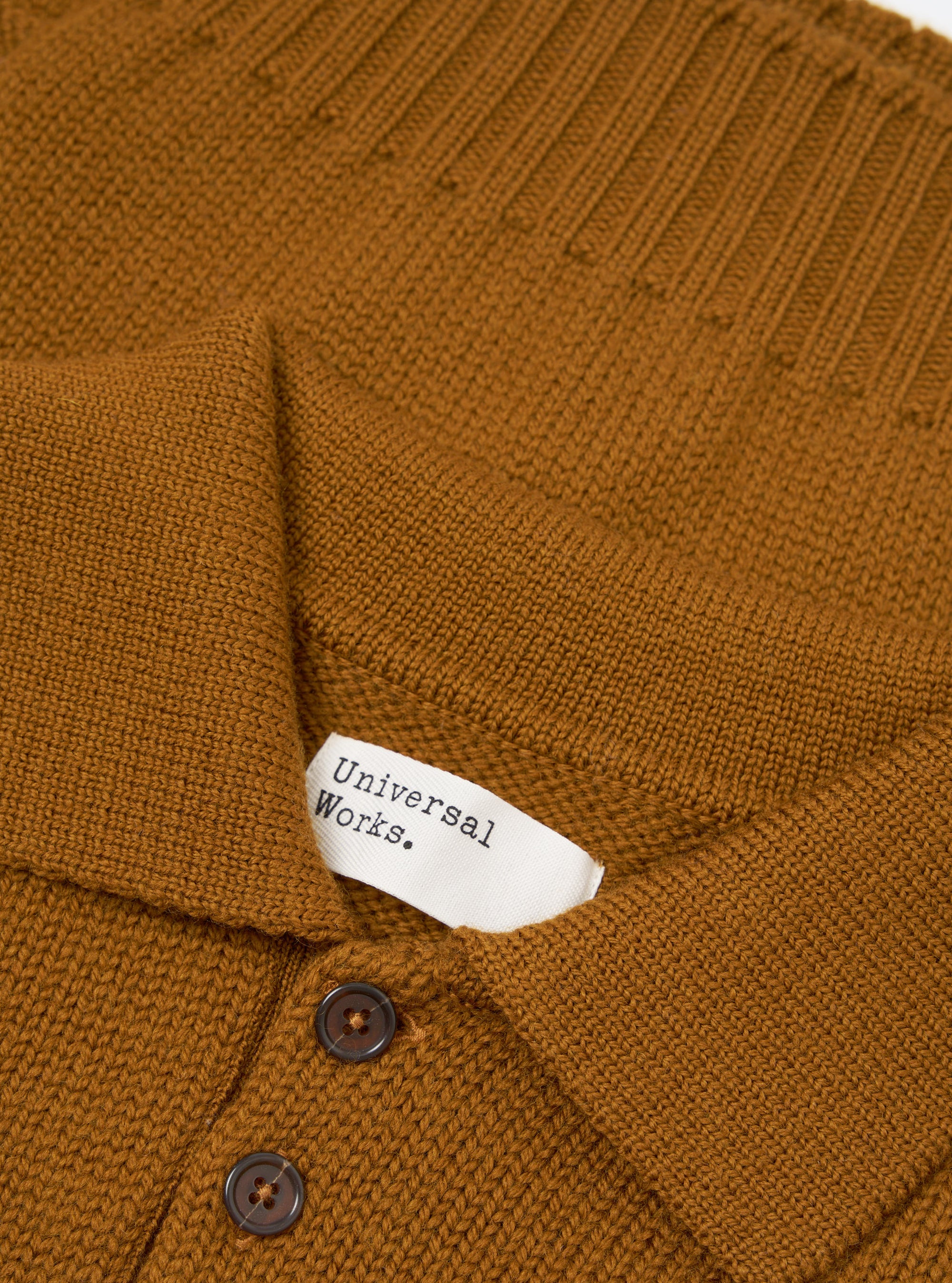 Close-up flat lay of a gold Shetland knit Curtis sweater by Universal Works. The spread collar and upper button placket are shown, with two dark horn-effect buttons visible. A white woven brand label reads "Universal Works." The knit texture varies between plain and ribbed sections across the body.