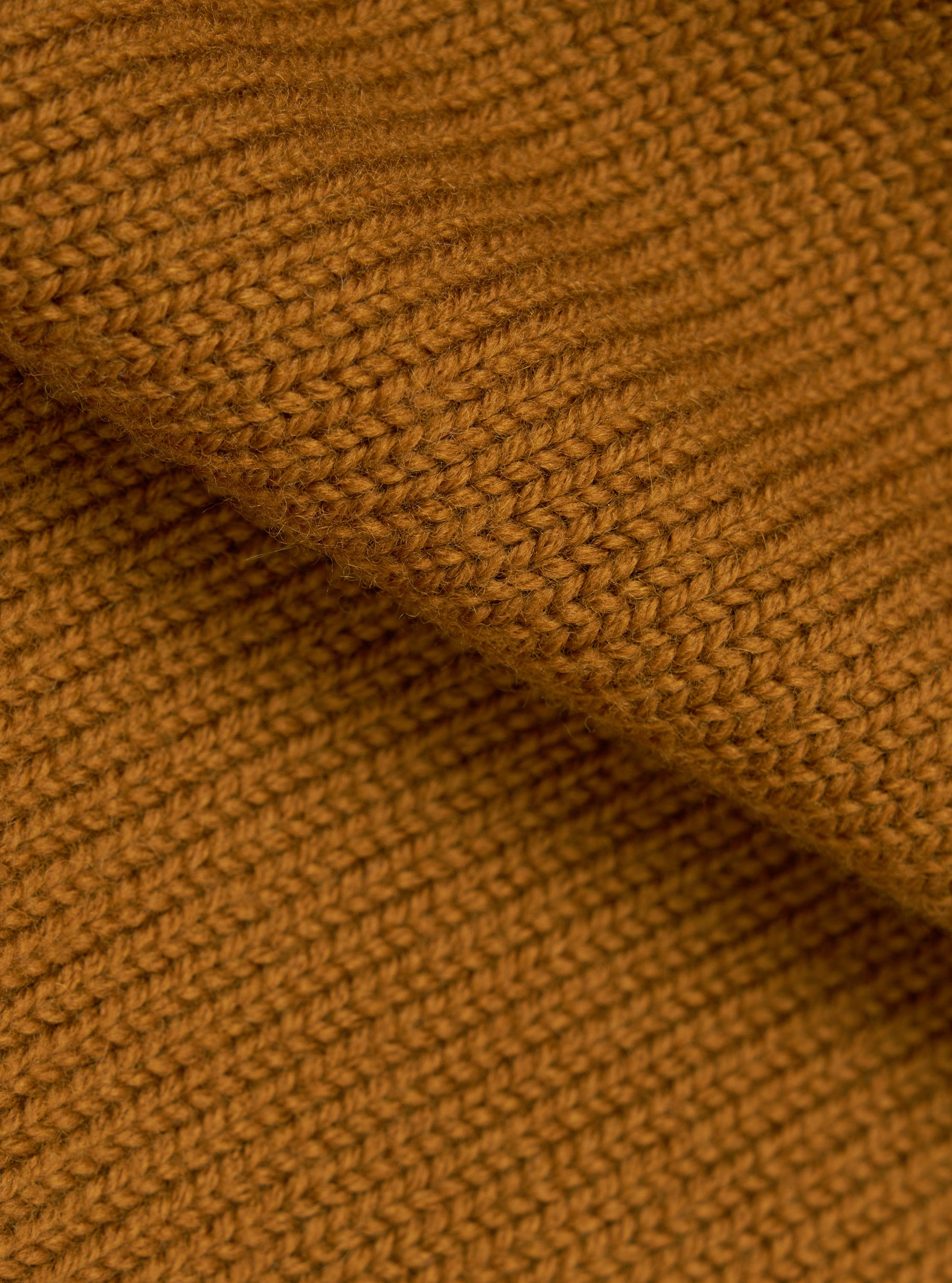 Close-up detail shot of the gold Shetland knit fabric on the Curtis sweater. The chunky knit texture is clearly visible, showing individual yarn loops in a warm amber-gold tone. A folded section runs diagonally across the frame, highlighting the fabric's depth and softness.