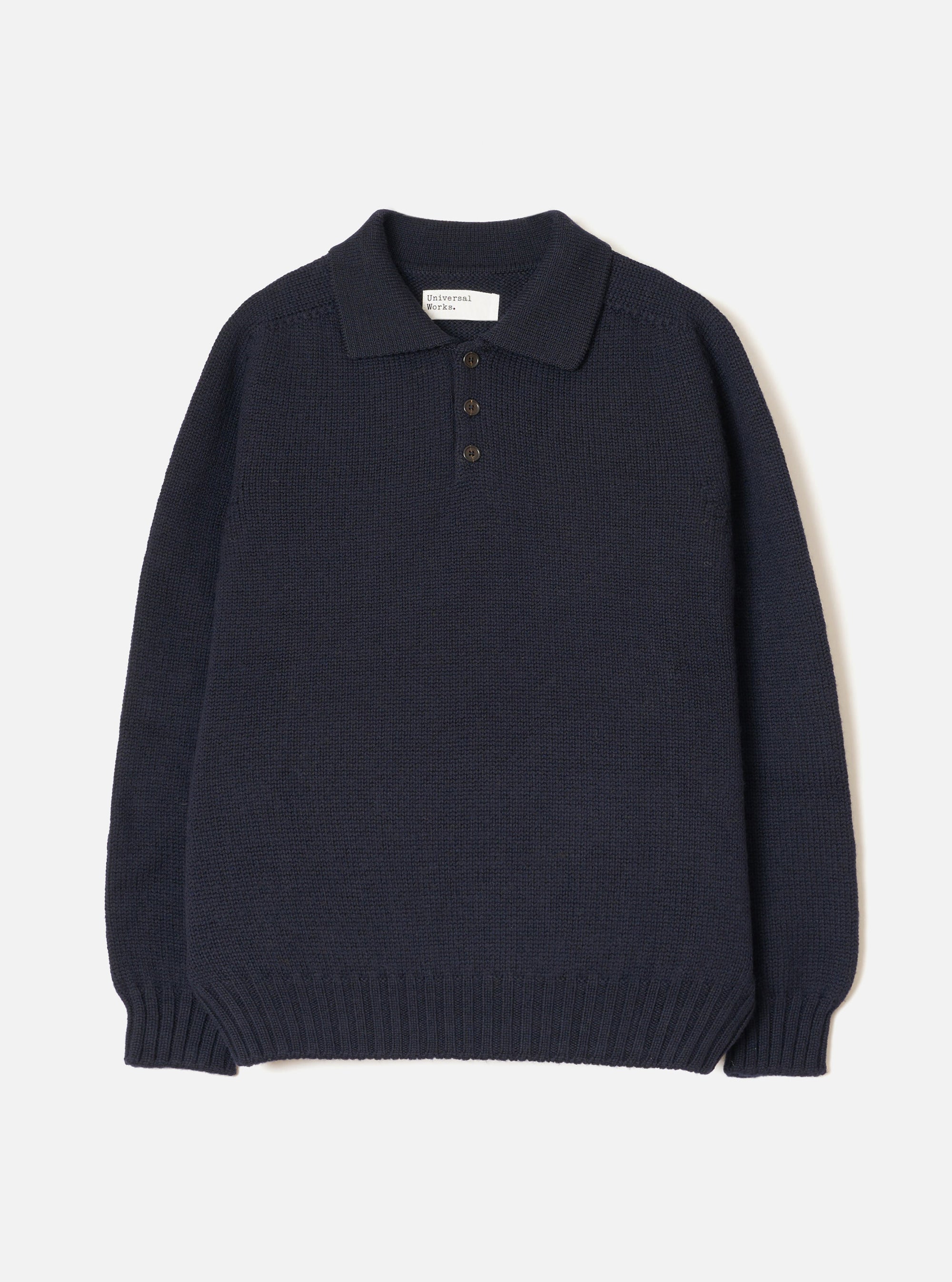 Navy Shetland knit polo sweater laid flat on a light grey seamless backdrop. Features a spread collar, three-button placket with dark buttons, chunky textured knit body, long sleeves, and a wide ribbed hem and cuffs. A Universal Works woven label is visible at the collar.