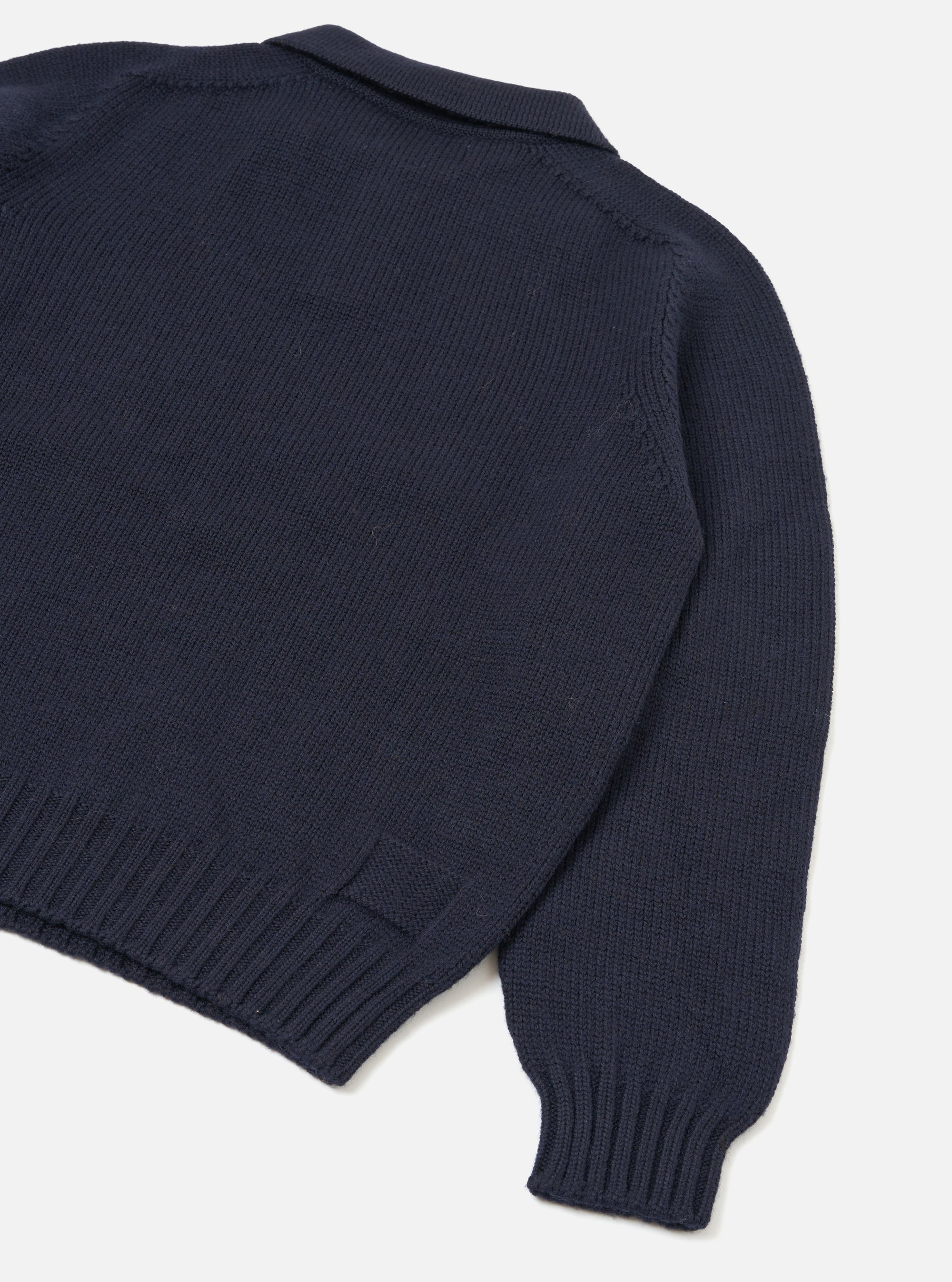 Navy Shetland knit polo sweater shot flat on a light grey seamless backdrop, angled to show the back and one sleeve. The body features a textured stocking-stitch knit with ribbed side panels, ribbed hem and ribbed cuffs. A spread collar is visible at the top. The sleeve seam shows a neat linking stitch detail.