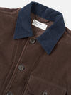 Close-up flat shot of the Universal Works cacao corduroy utility jacket on a light grey backdrop. The wide-wale cord body is a warm brown, contrasted by a navy corduroy collar. Dark four-hole buttons fasten the front placket. A chest patch pocket is visible at the lower frame. The branded woven label sits at the neckline.