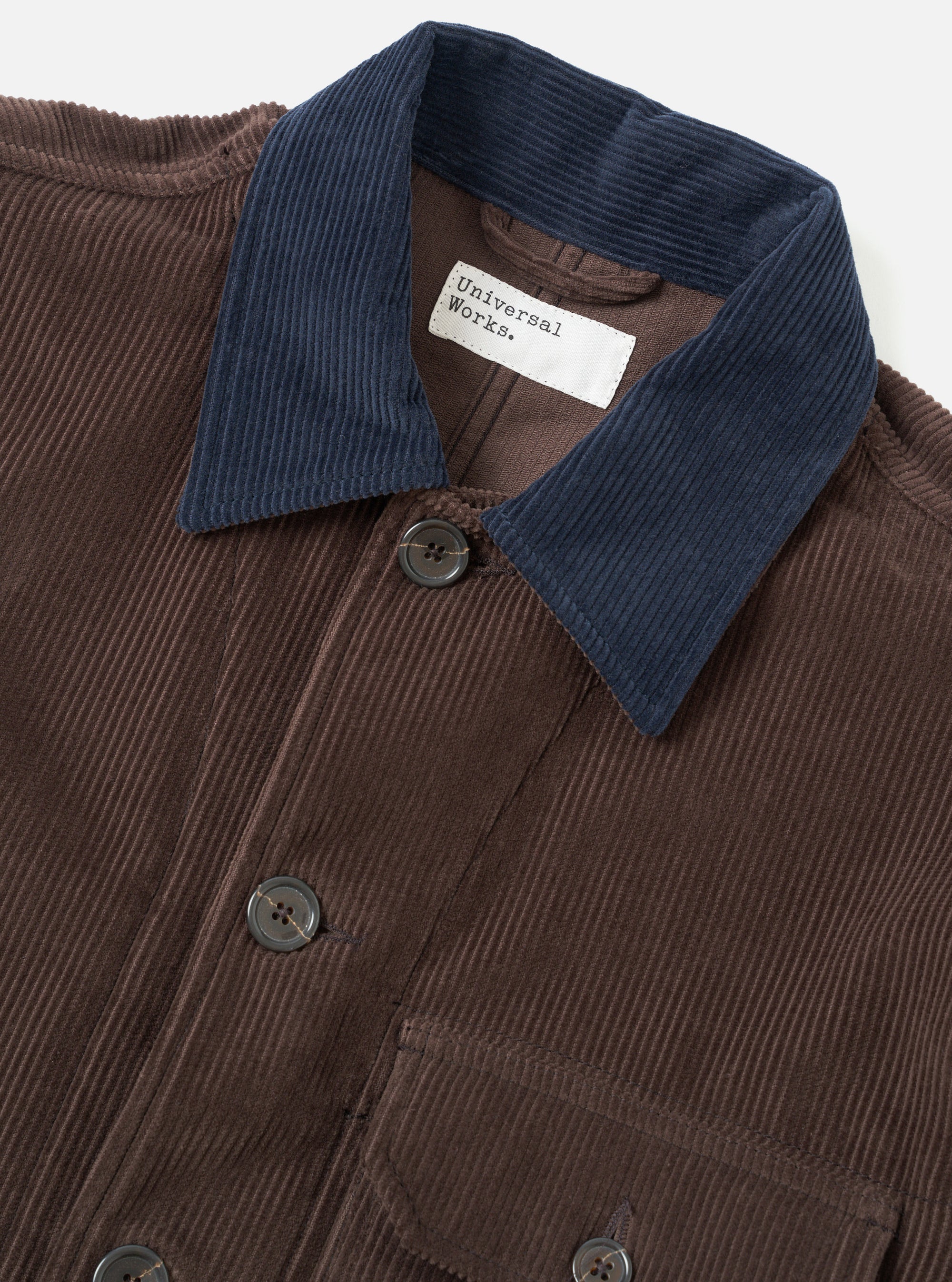 Close-up flat shot of the Universal Works cacao corduroy utility jacket on a light grey backdrop. The wide-wale cord body is a warm brown, contrasted by a navy corduroy collar. Dark four-hole buttons fasten the front placket. A chest patch pocket is visible at the lower frame. The branded woven label sits at the neckline.