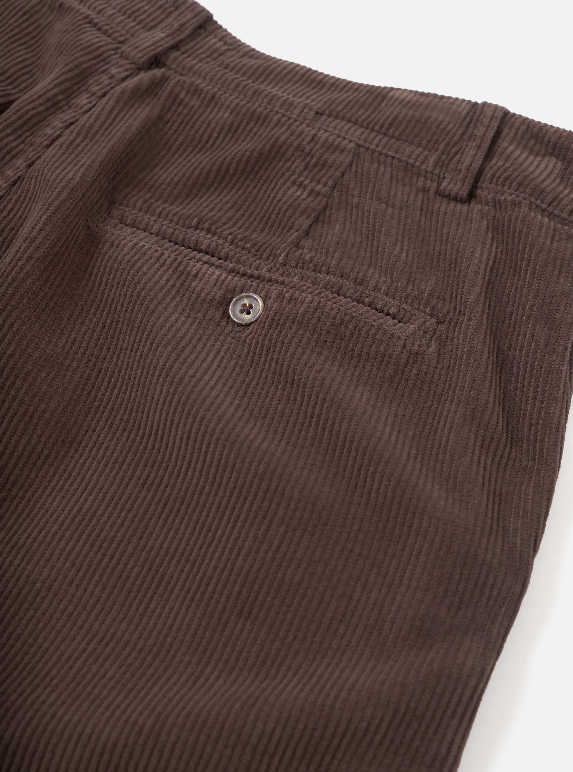 Close-up detail of cacao brown corduroy military chino trousers on a white studio backdrop. The wide-wale cord texture is clearly visible. A single welt back pocket with a four-hole button fastening is shown, alongside a belt loop and waistband at the top of the frame.
