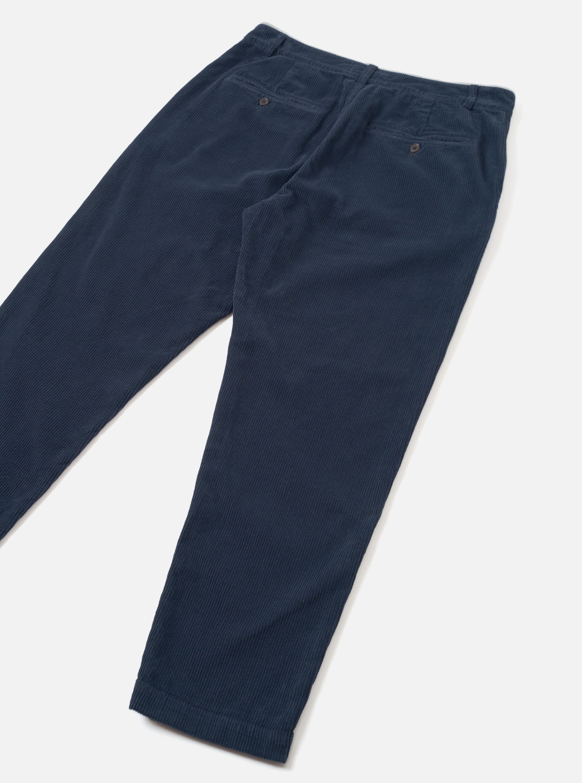 Dark navy corduroy military chinos laid flat on a light grey surface, shot at an angle. Fine wale cord texture is visible across both legs. The waistband features belt loops and a buttoned welt back pocket. The tapered legs end in plain, unfolded hems.