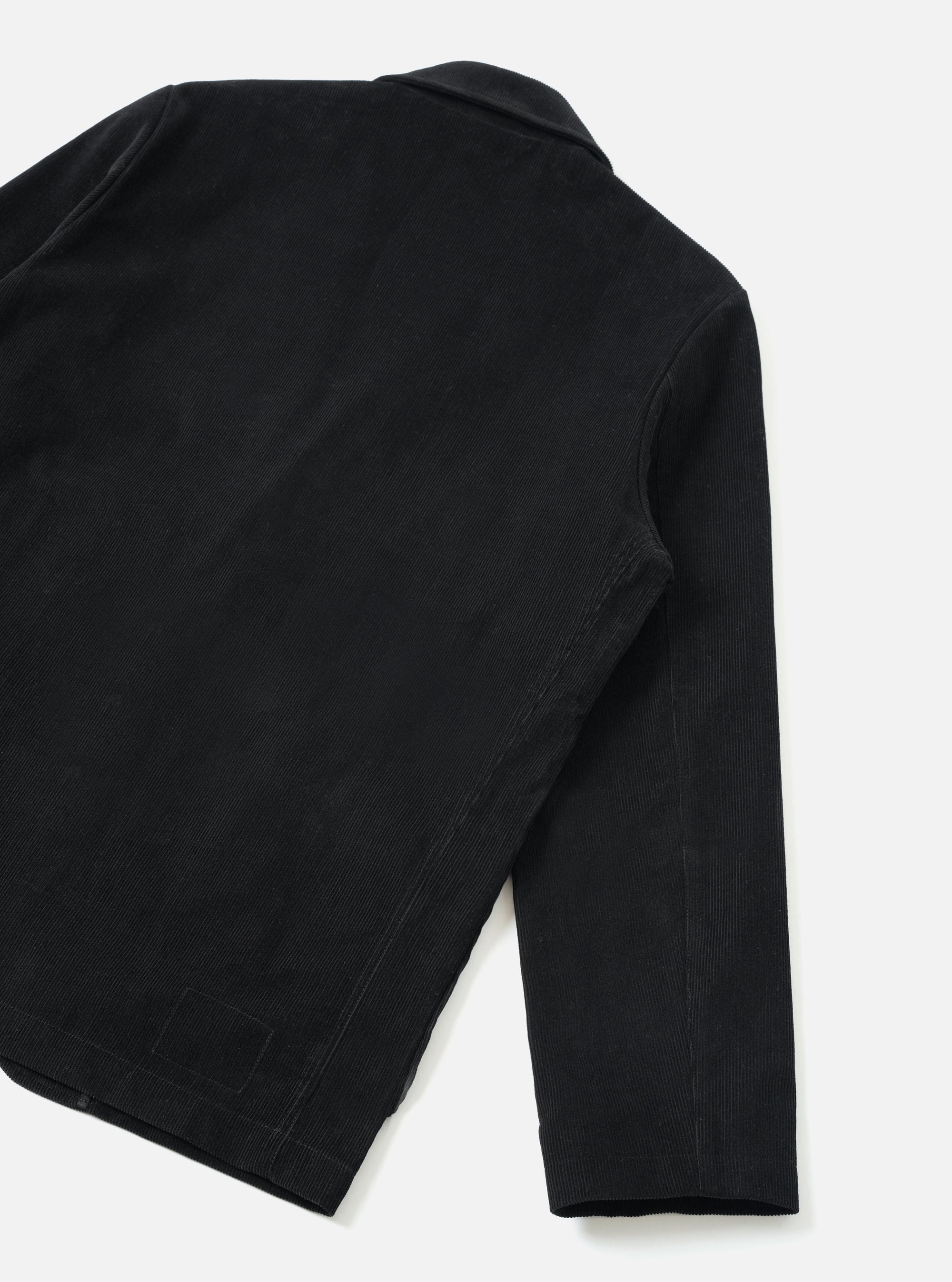 Flat lay detail shot of the black corduroy two-button jacket on a light grey seamless backdrop, showing the back and one sleeve. Fine wale corduroy texture is visible across the body and sleeve. Topstitched seams run along the armhole and hem. A small woven label patch sits at the lower back hem.