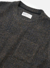 Close-up flat lay of a Universal Works navy Deca Light Knit Sleeveless Crew on a white studio surface. The marl fabric blends navy, brown and blue tones in a fine knit texture. A ribbed crew neckline is visible with a dark inner tape, and a woven Universal Works label sits at the front neck.