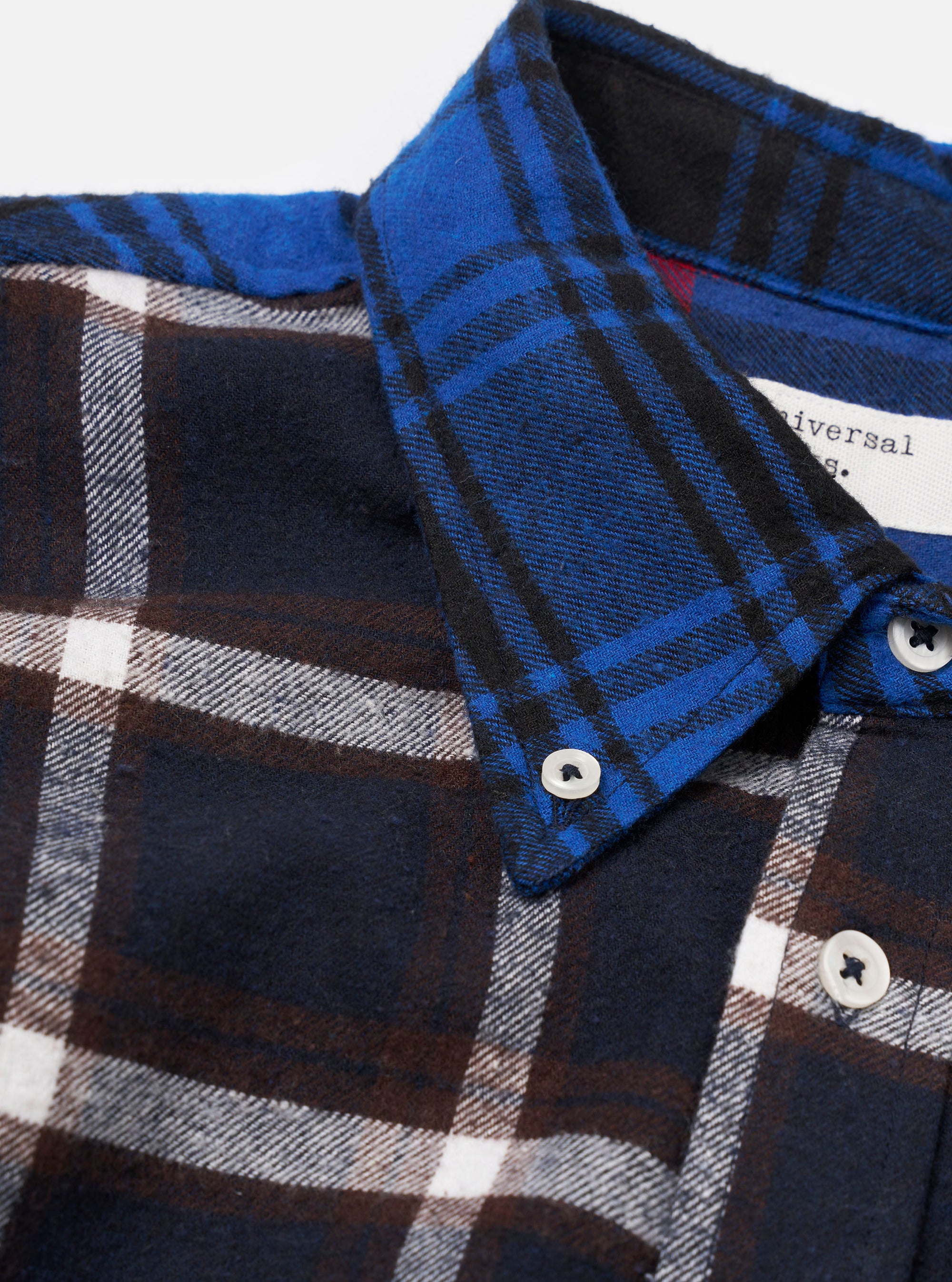 Close-up of a blue patchwork check flannel shirt on a white studio backdrop. The collar and button placket are shown in detail, with the collar cut from a bold blue and black check contrasting the body's navy, brown and white large-scale check. White buttons fasten the collar and placket. A woven Universal Works label is visible at the neckline.