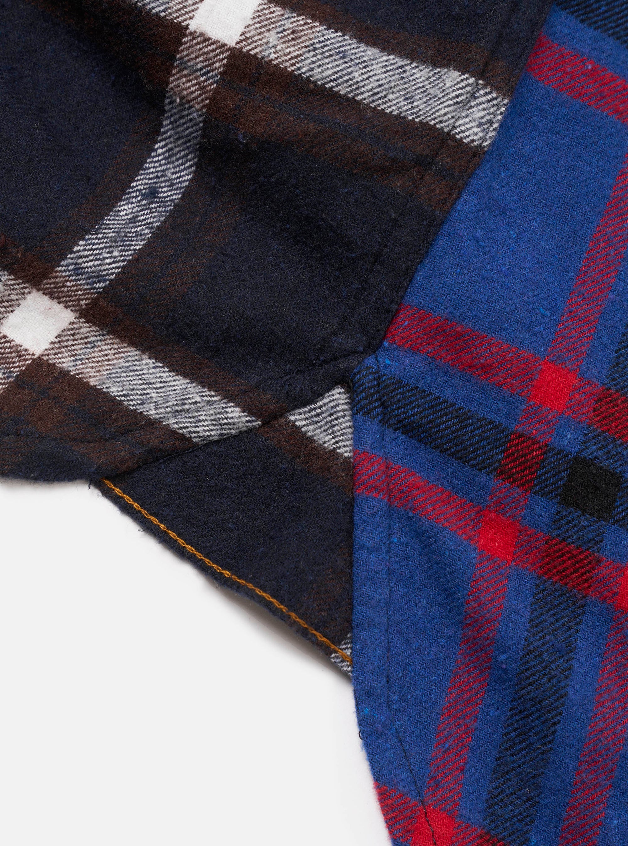 Close-up flat lay of a blue patchwork checks shirt on a white surface. Two contrasting tartan panels are visible: a dark navy, brown and white check on the left, and a bright blue, red and black tartan on the right. A seam joins the panels with visible mustard yellow stitching. Soft, brushed flannel texture throughout.
