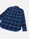 Navy and blue brushed flannel check shirt laid flat on a light grey seamless backdrop, showing the back view. Bold tartan check pattern in blue and black covers the body and long sleeves. A centre back pleat, curved hem, small woven label at the lower hem, and two white buttons at the cuff are visible.