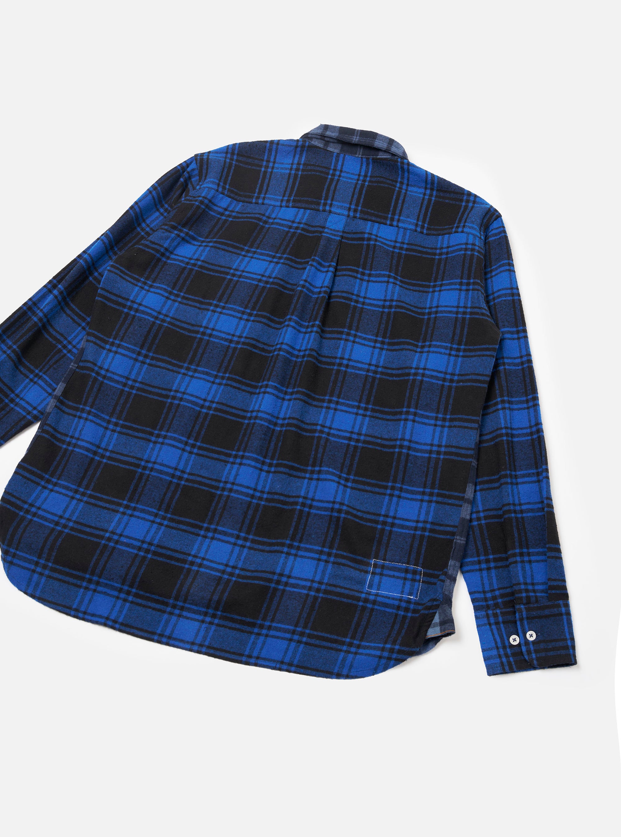 Navy and blue brushed flannel check shirt laid flat on a light grey seamless backdrop, showing the back view. Bold tartan check pattern in blue and black covers the body and long sleeves. A centre back pleat, curved hem, small woven label at the lower hem, and two white buttons at the cuff are visible.