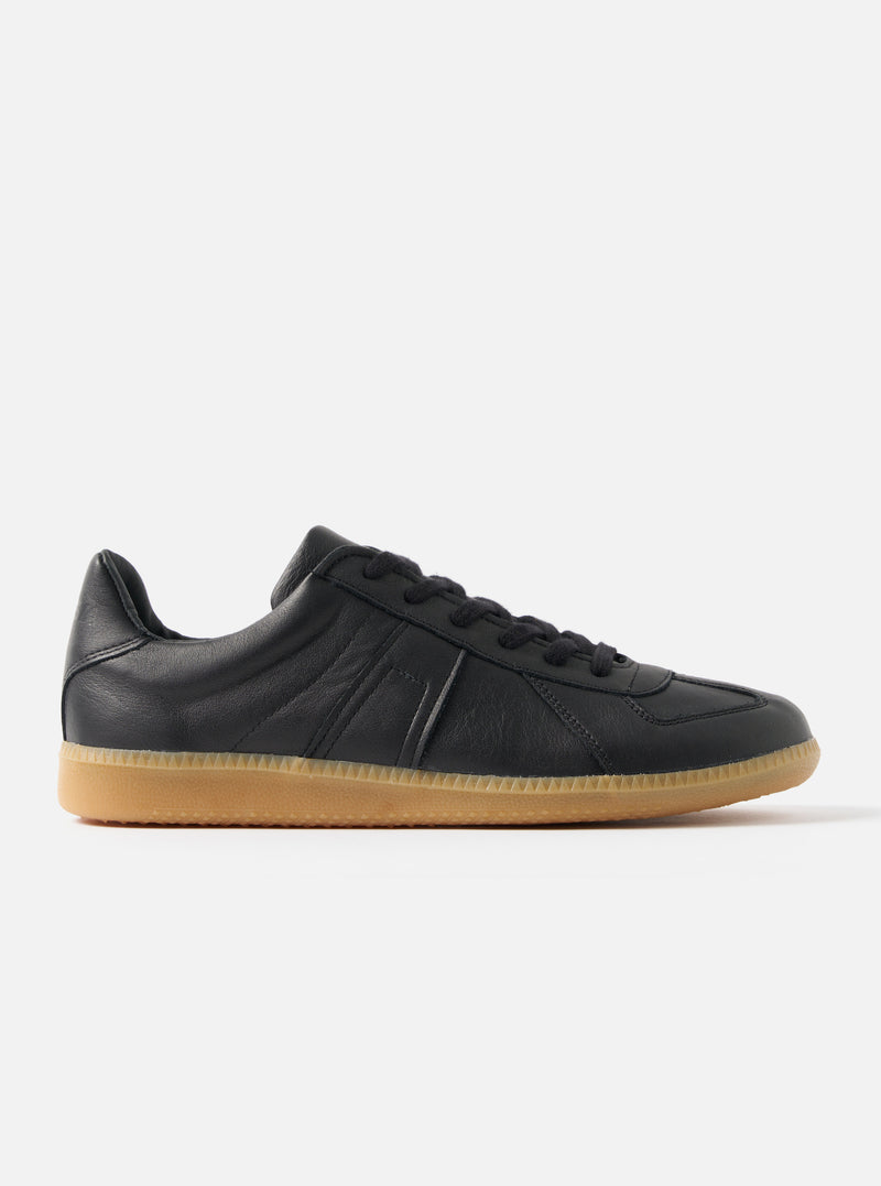 Side view of the Novesta for Universal Works Black Leather German Army Trainer on a white background. The shoe has a black leather upper and a gum-colored sole.