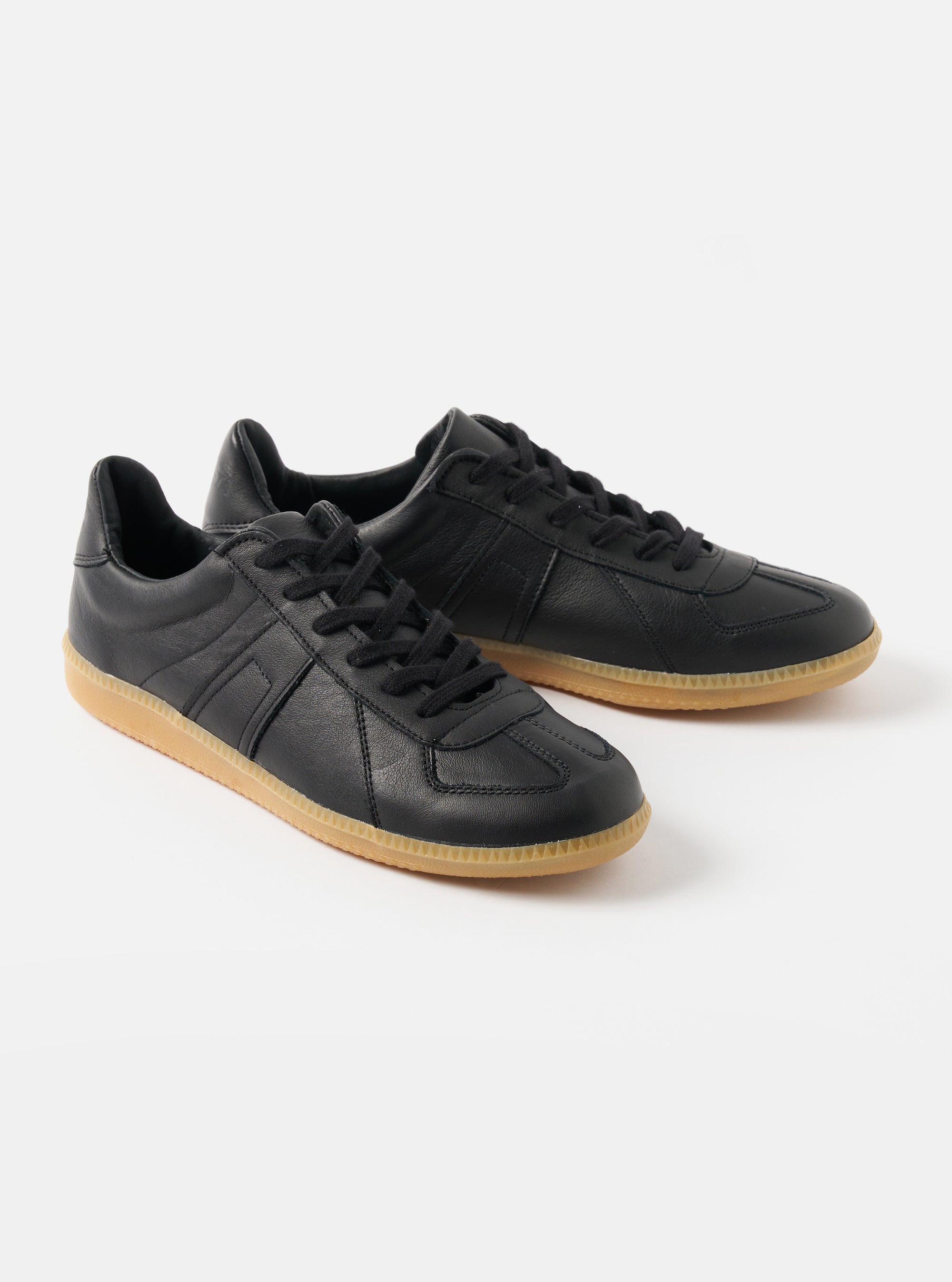 Studio shot of Novesta for Universal Works Black Leather German Army Trainer shoes. The trainers have black leather uppers and tan rubber soles, set against a white background.