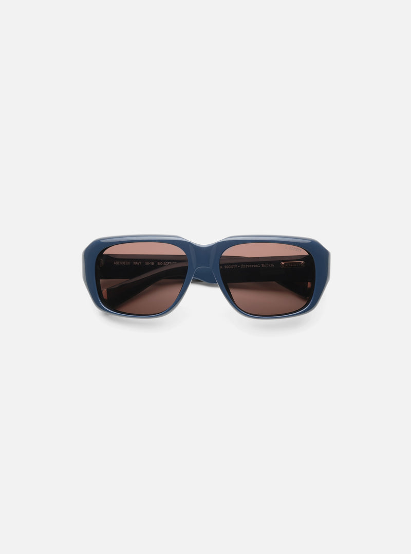 Close-up of A. SOCIETY x Universal Works Navy Aberdeen Sunglasses on a white surface. The frames are navy, and the lenses are brown.
