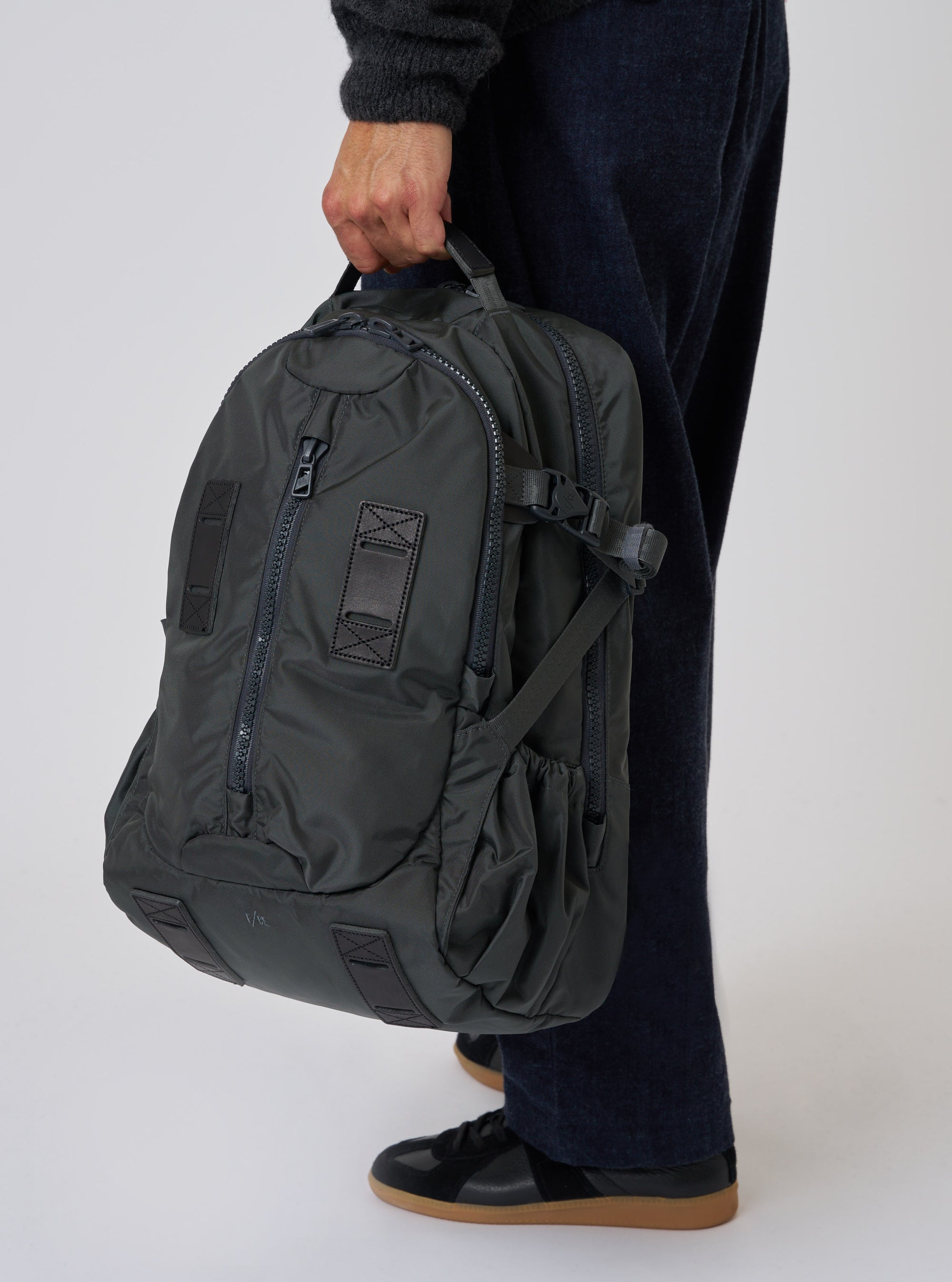 A person holds the F/CE.® Grey Recycled Nylon 420 re/cor Travel Backpack. The grey nylon backpack is held against a white backdrop, with a person wearing dark blue pants and black sneakers.