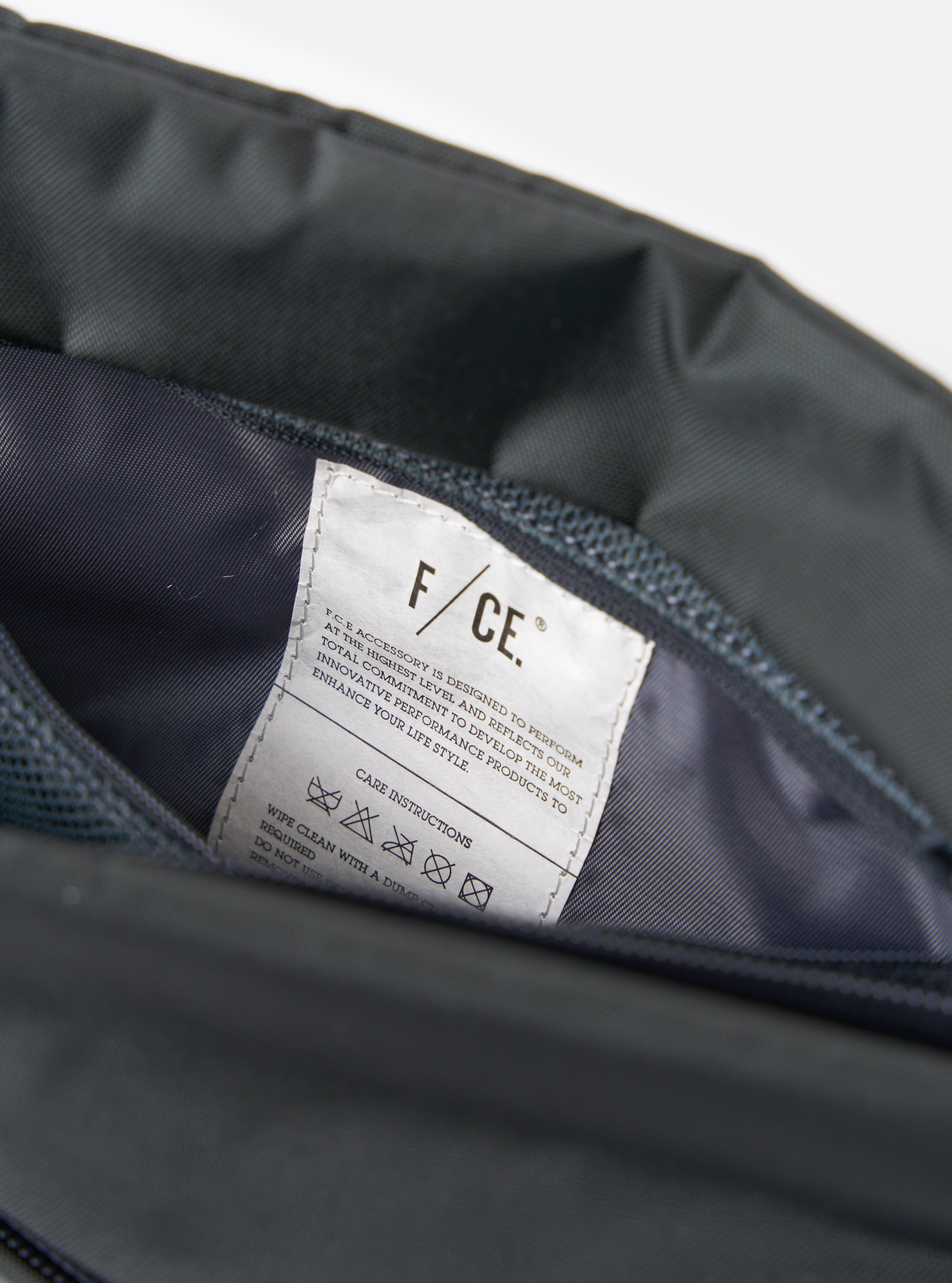 Close-up of the inside of the F/CE.® Grey 420 re/cor Sacoche, showing the grey material and a white tag with care instructions. Shot on a white background.