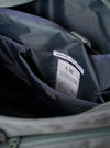 The F/CE. Foliage Green Recycled Nylon 420 re/cor Courier Shoulder Bag is worn by a model against a plain, light grey background. The model carries the bag across the body, highlighting its large, messenger-style silhouette and wide, adjustable shoulder strap. The foliage green recycled nylon fabric, front pocket details, and the way the bag sits snugly against the model's back are the prominent features of the image.