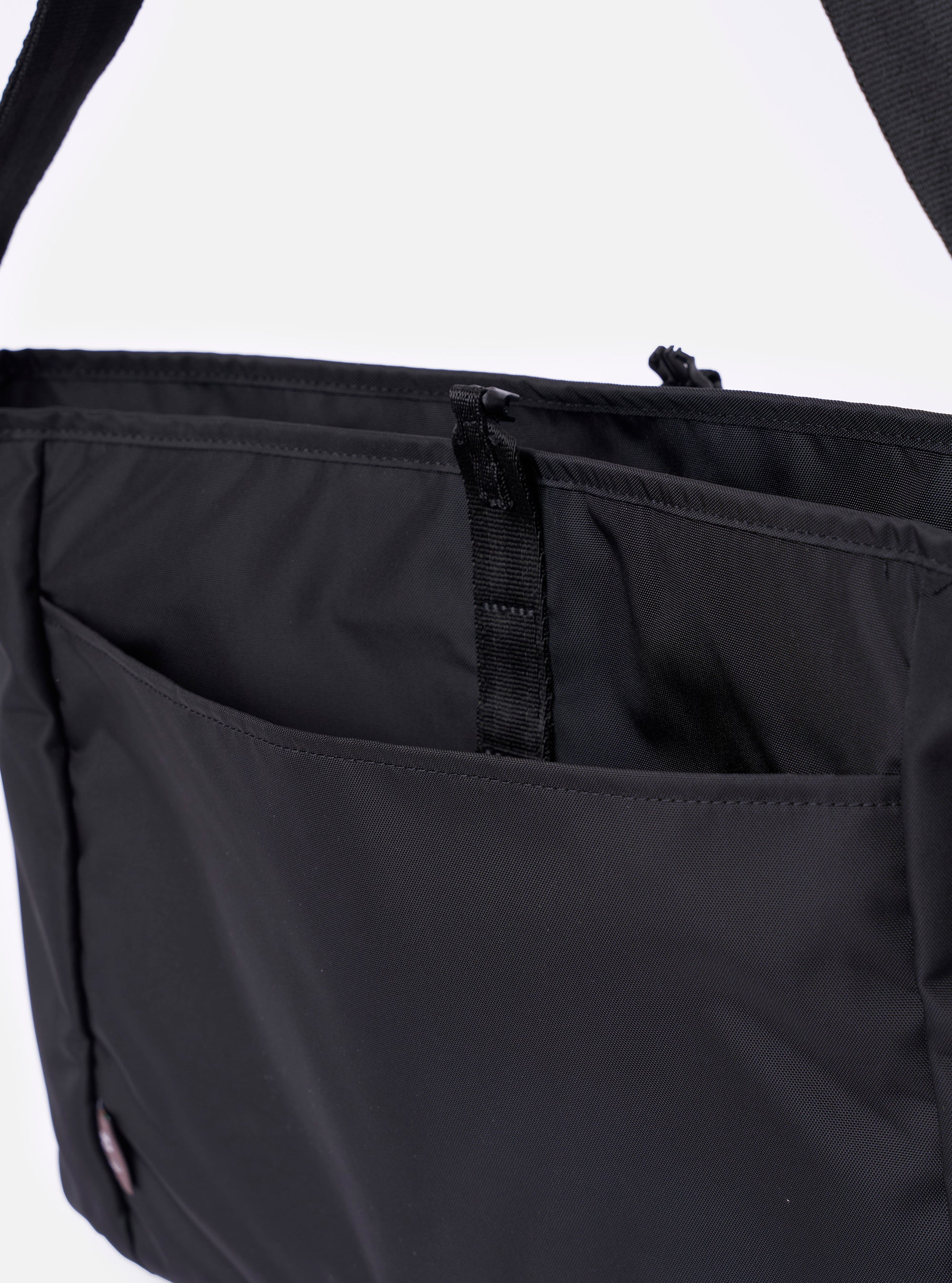 The F/CE. Black Recycled Nylon 420 re/cor Courier Shoulder Bag is shown from a side-profile angle as a flat lay against a plain white background. The image highlights the bag's depth and the technical construction of the adjustable messenger-style shoulder strap, which features custom-ordered nylon tape and a metal adjustment mechanism. The black recycled nylon fabric has a subtle matte-shiny finish.
