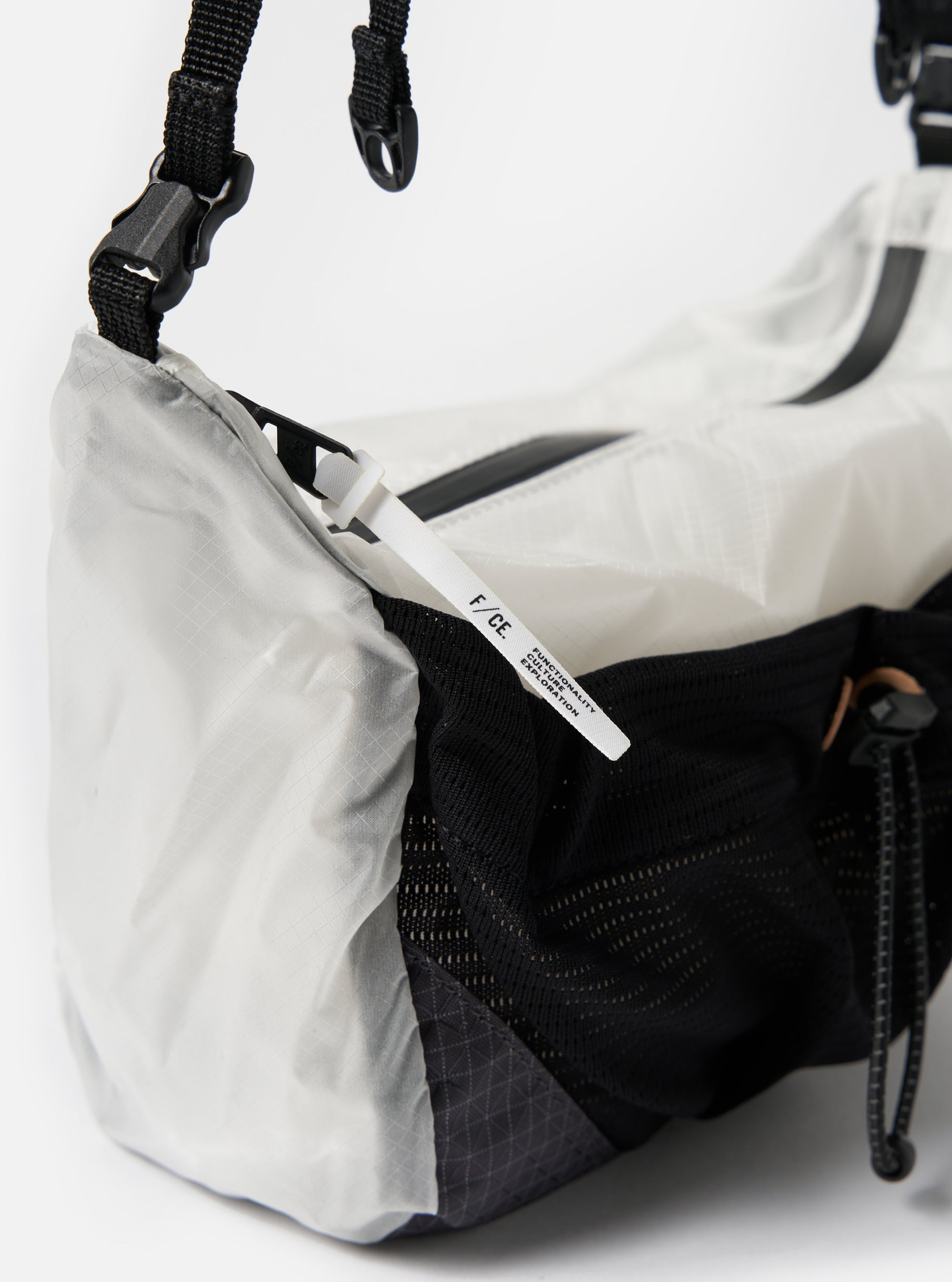 Close-up of the F/CE.® White Nylon Cordura Sil Sacoche against a white background. Features black straps and mesh, and a white tag. Shows the bag's texture and design details.